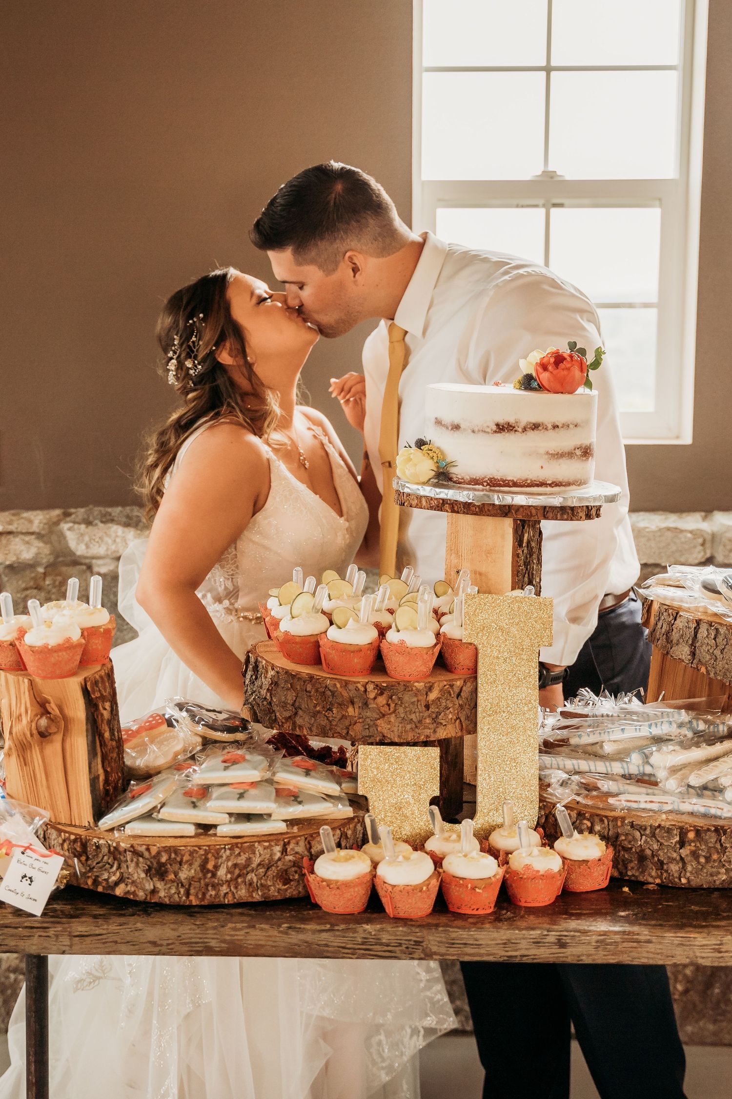 Bride and groom kissing at rustic wedding dessert table with tiered cake and cupcakes.