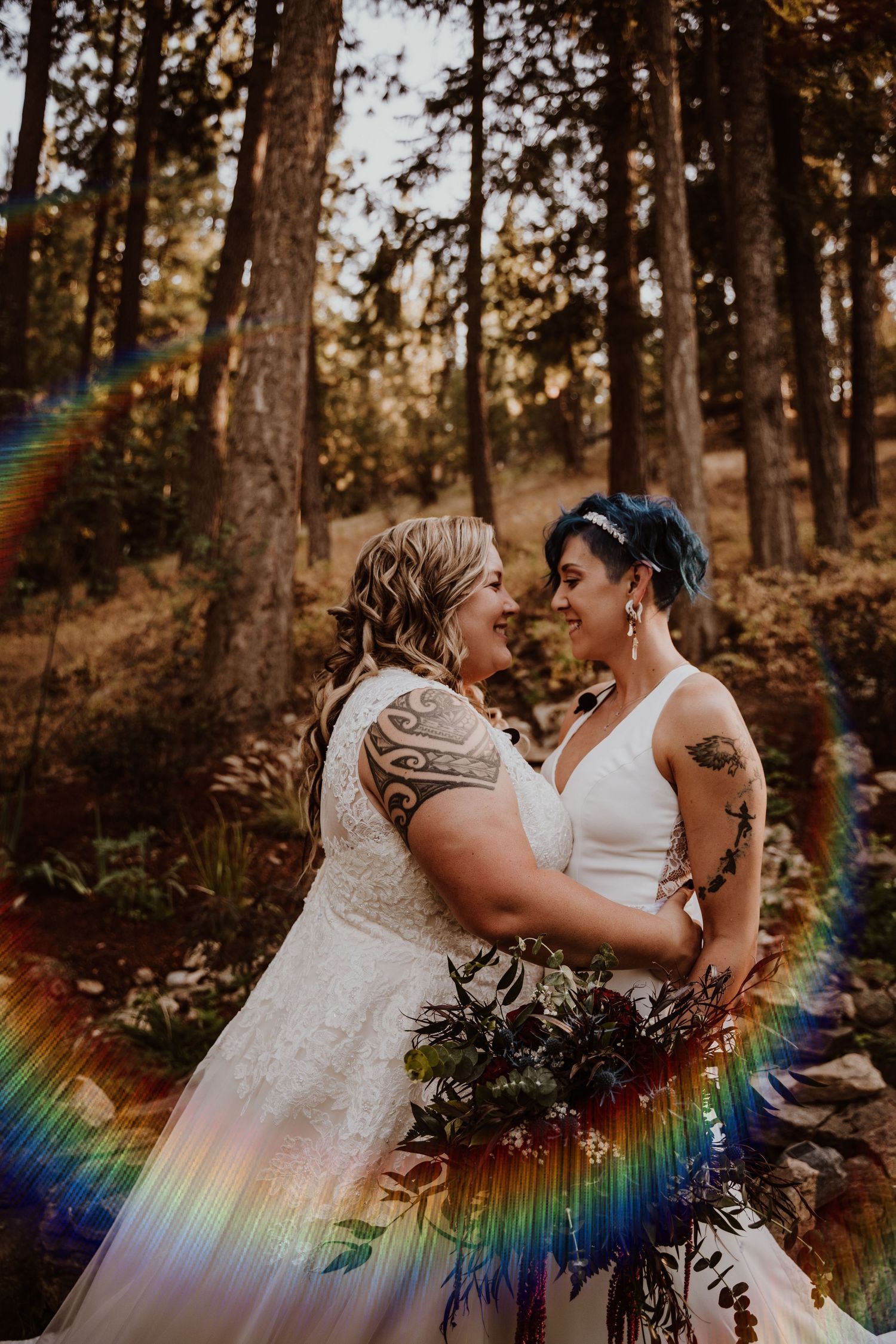 Two brides in white wedding gowns embracing in forest with rainbow light prism effect.