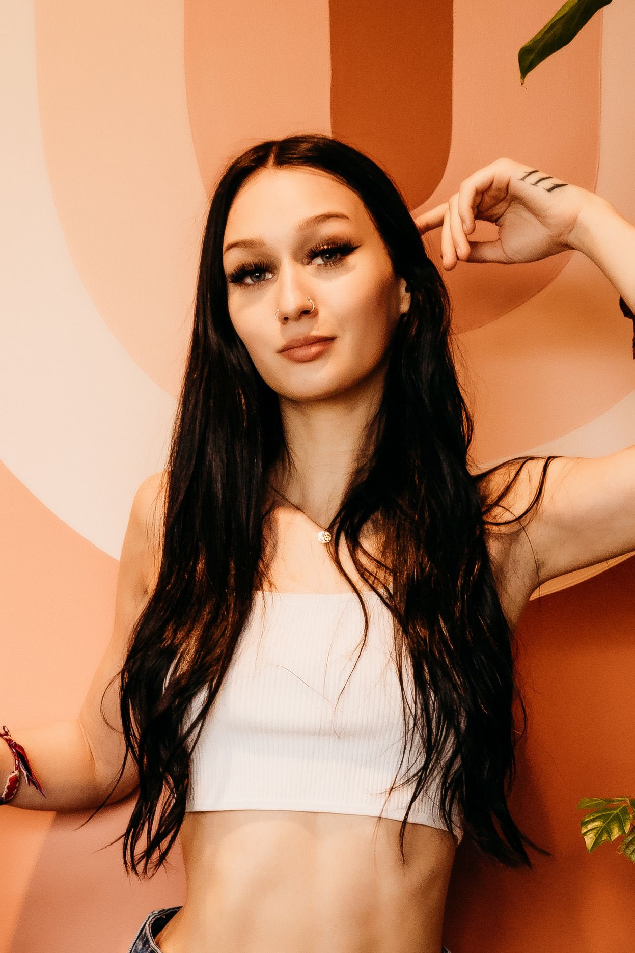 Young woman with long dark hair in white crop top posing against a peach-toned abstract background.