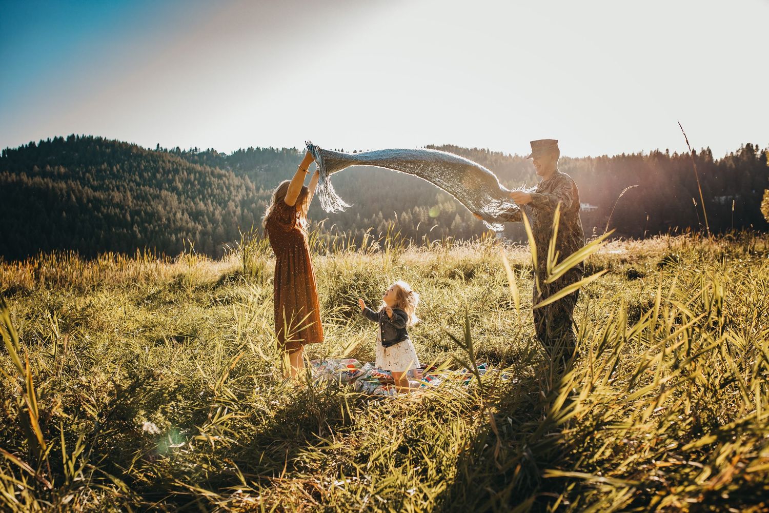 Family of three playing with a giant soap bubble in a sunlit mountain meadow with forest in the background.