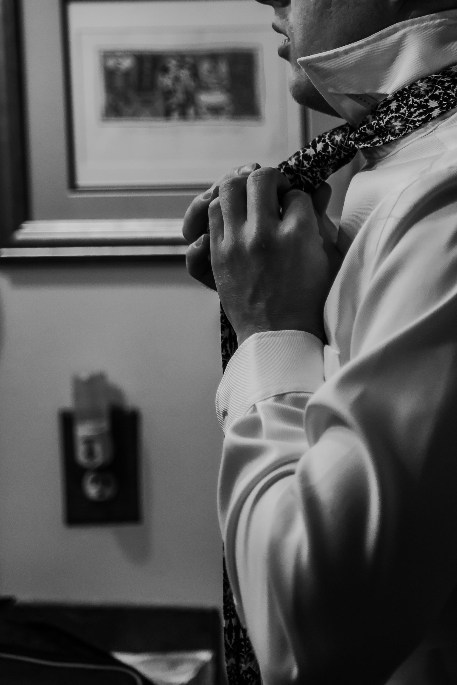 Groom adjusts patterned tie while getting ready in black and white wedding photo.