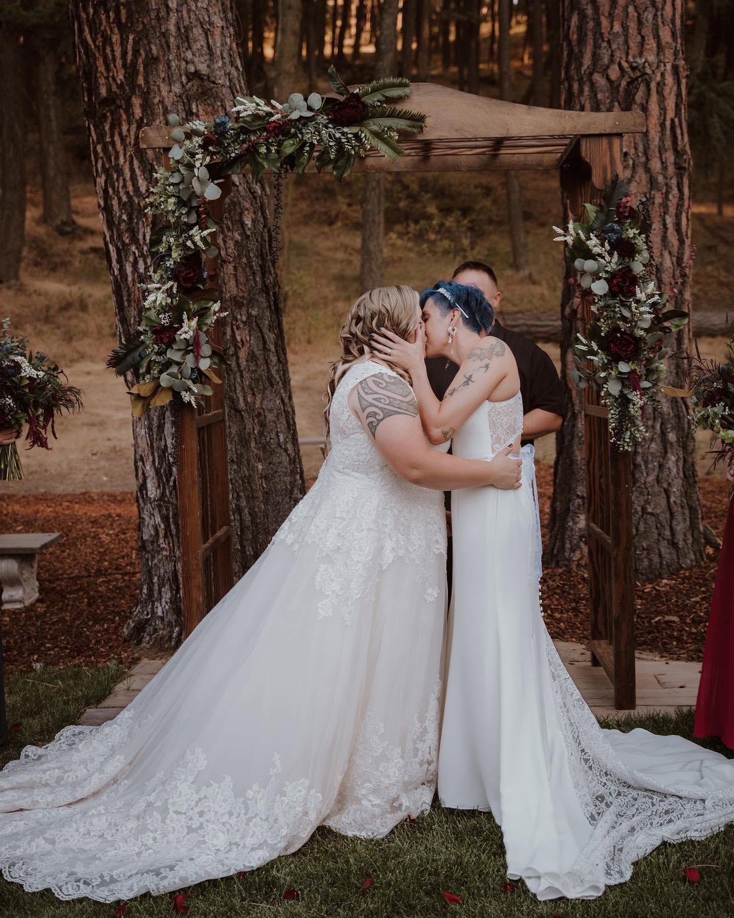 Two brides share their first kiss at a rustic wooden arch adorned with lush greenery and burgundy florals.