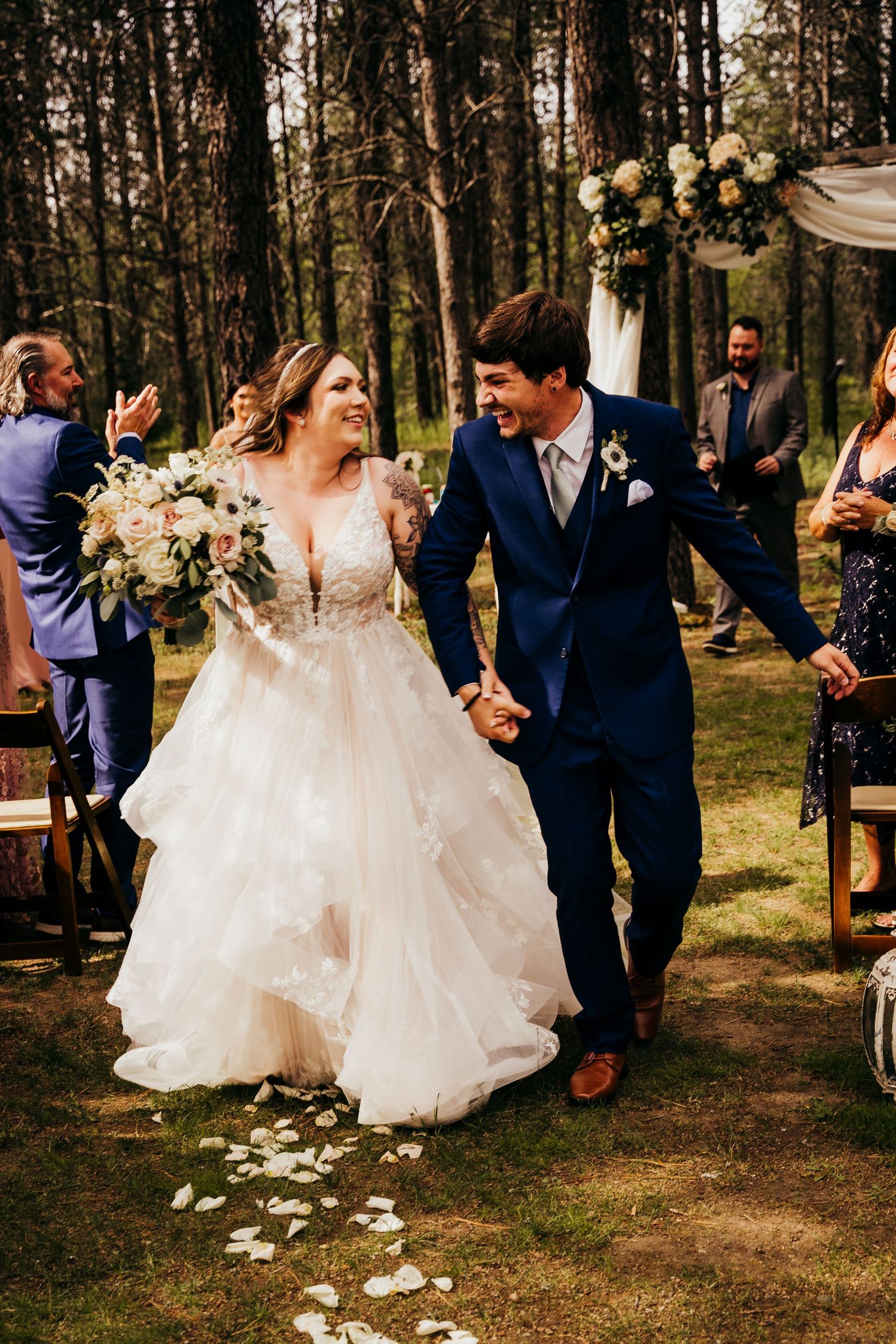 Radiant newlyweds holding hands and laughing while walking down the aisle in pine forest.