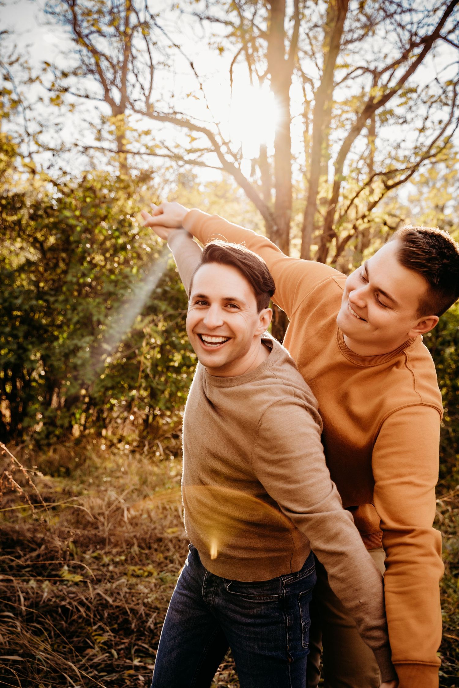 Two men laugh and play piggyback in a sunlit autumn forest during an engagement session.