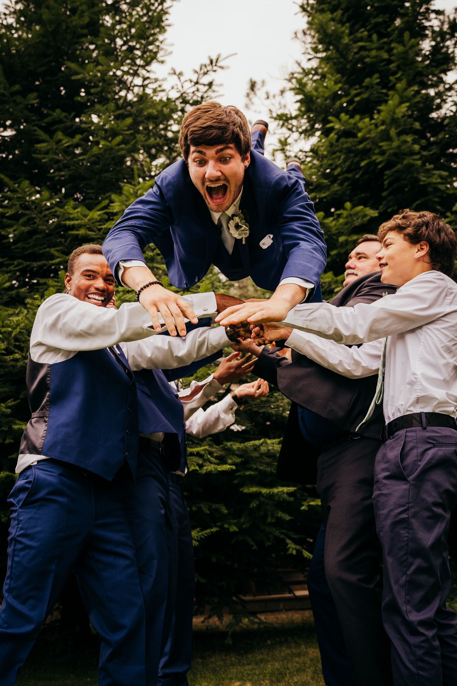 Groom in a blue suit being lifted by groomsmen outdoors near evergreen trees at a wedding celebration.
