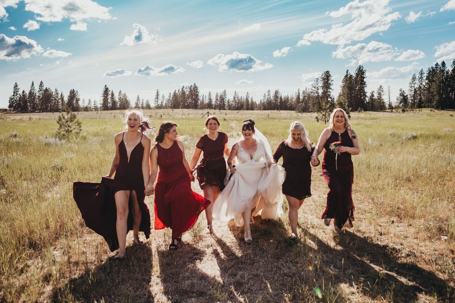 Bride and bridesmaids in black dresses walk together laughing through a sunny open meadow.