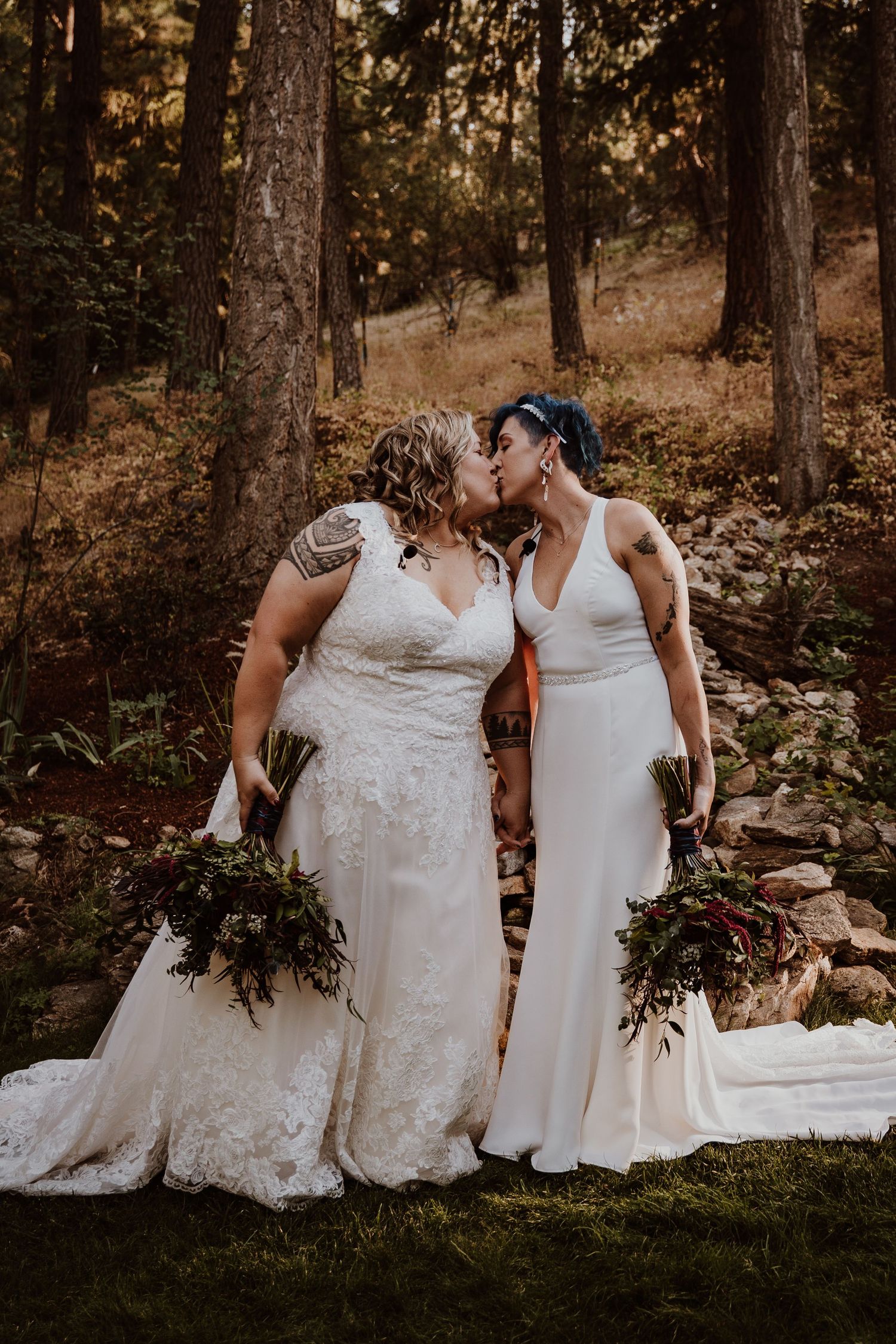 Two brides in white gowns share a romantic kiss while holding floral bouquets in a forest.