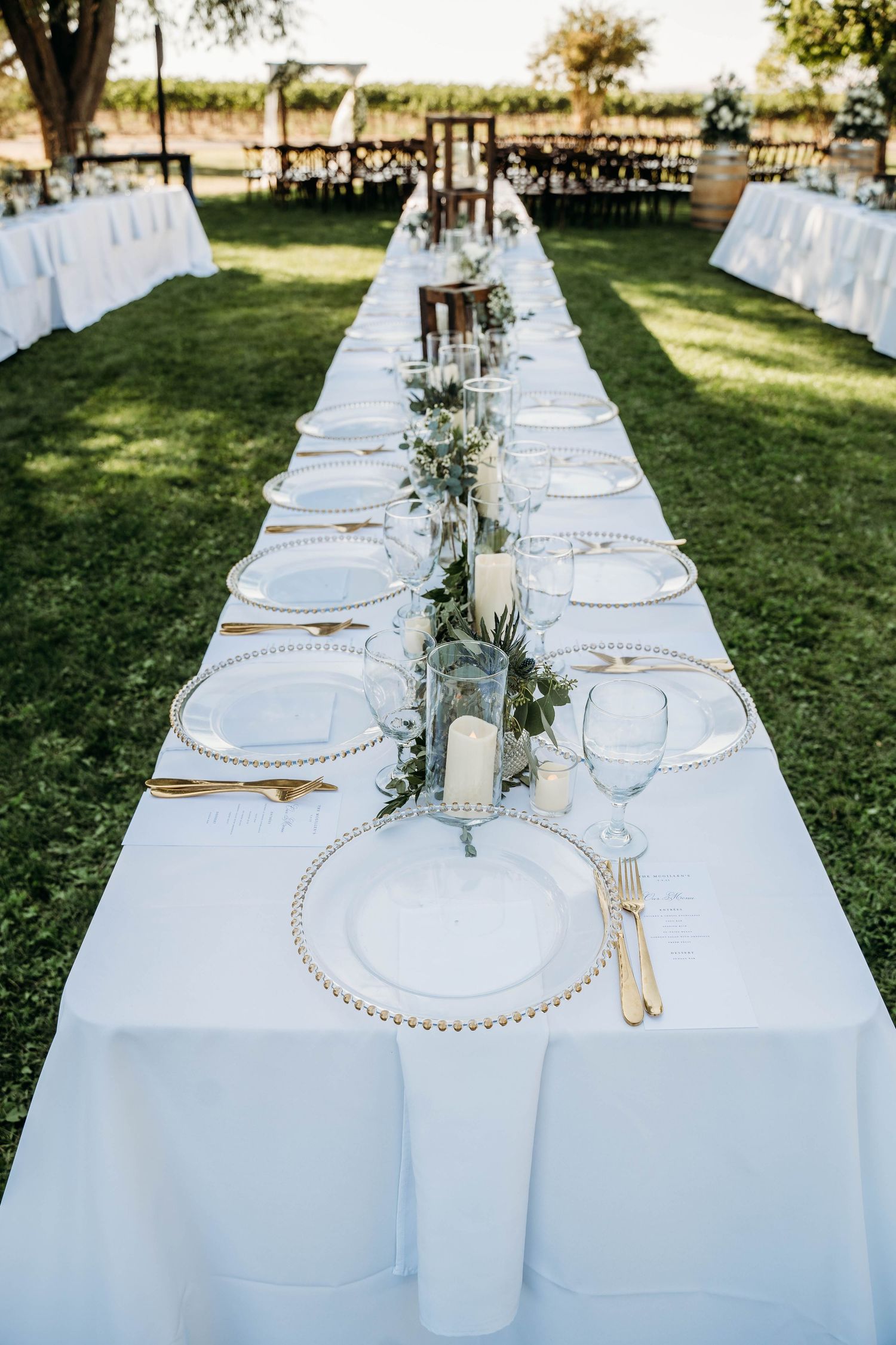 Elegant outdoor wedding reception with long white tablecloth, gold cutlery, greenery runner, and candles on green lawn.