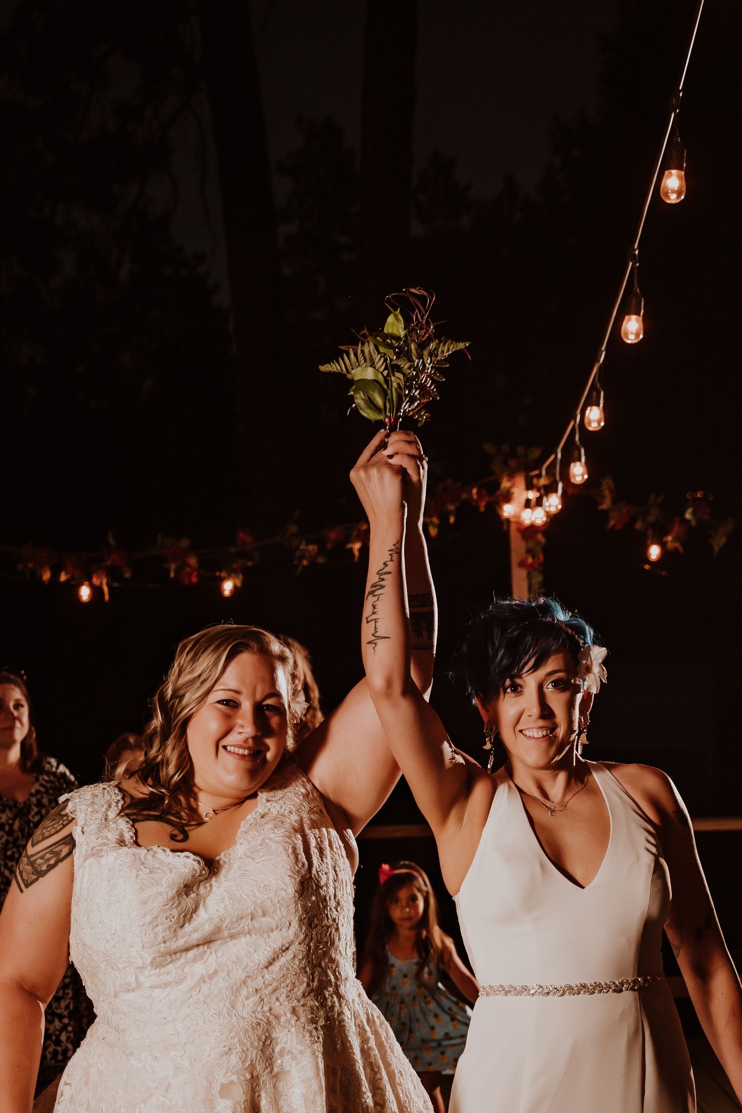 Two brides celebrate joyfully raising a bouquet under string lights at their wedding reception.