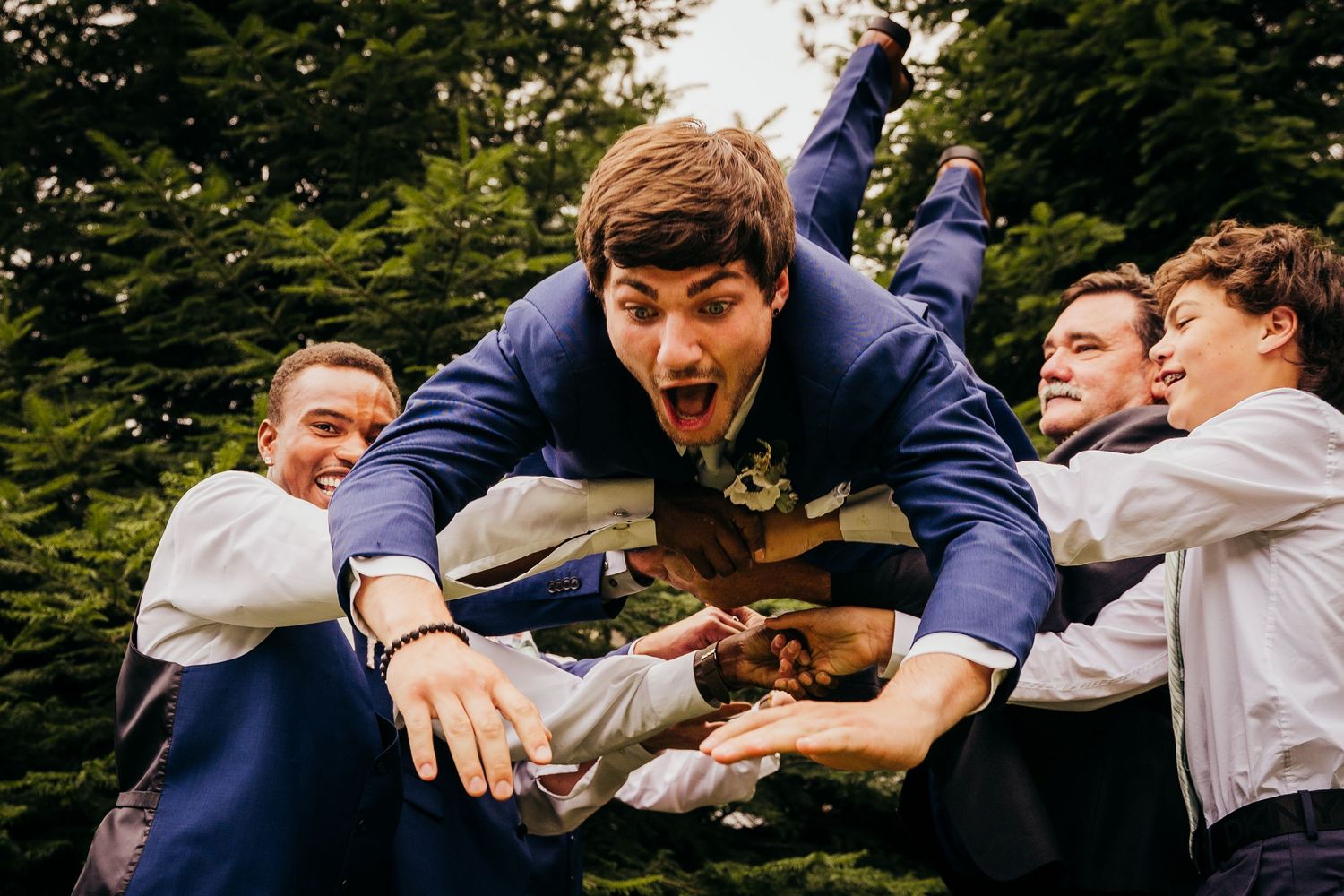 Excited groom being lifted and carried by laughing groomsmen in blue suits outdoors.