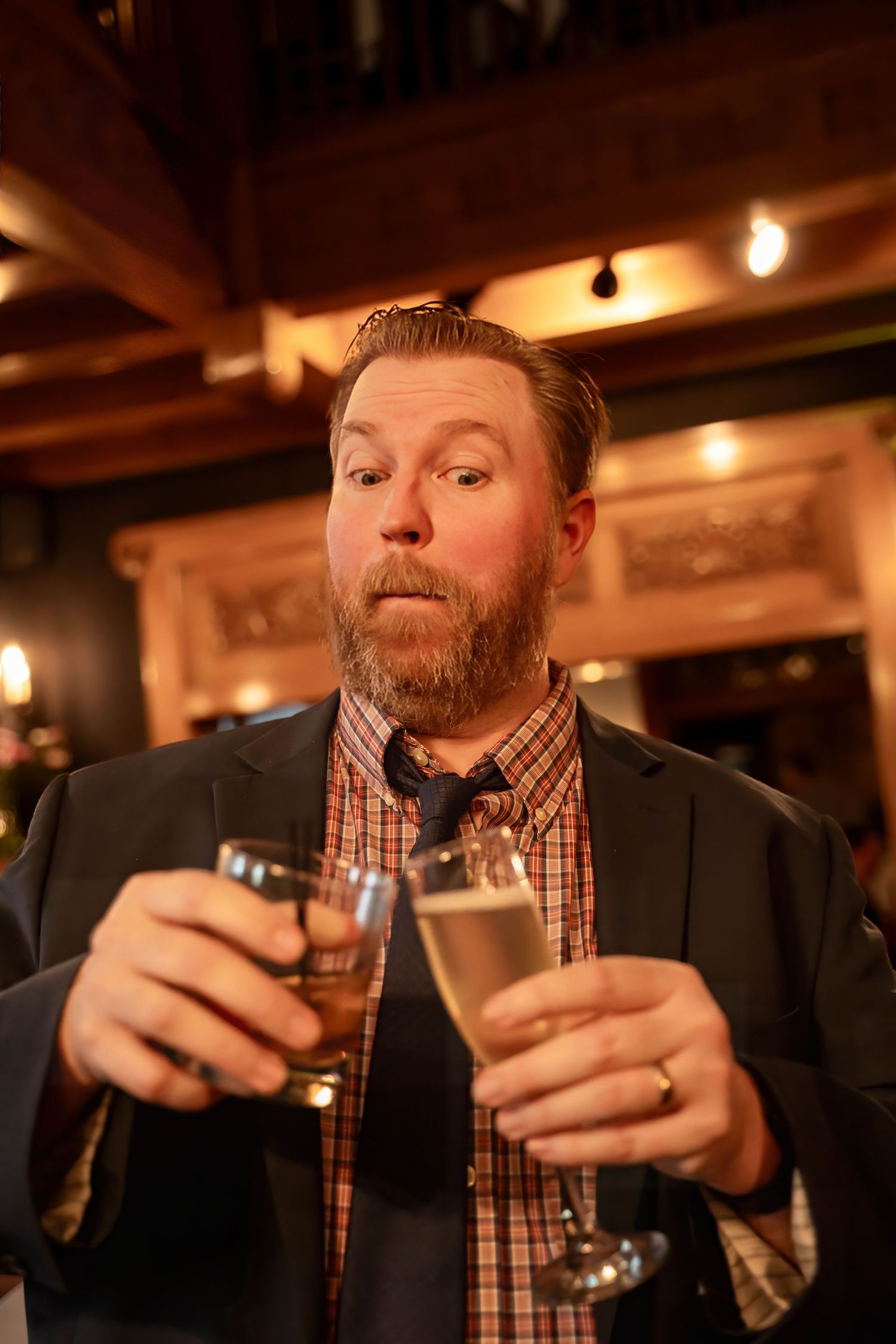 Bearded man in a blazer holds two champagne glasses with a surprised expression at a wedding reception.