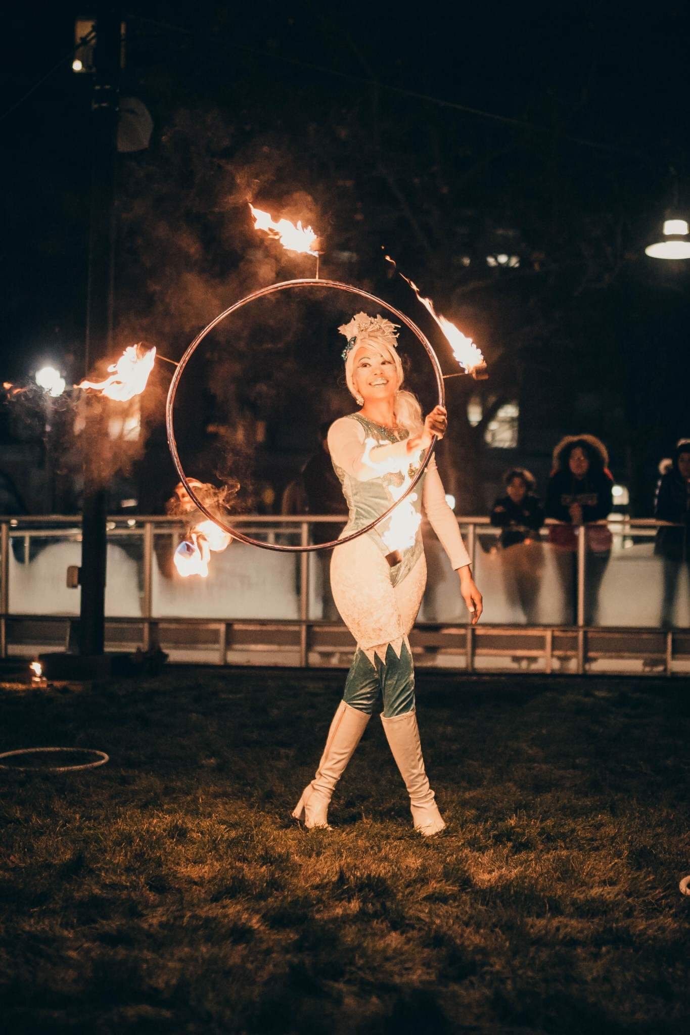 Female fire performer spinning flaming hoop during dramatic nighttime outdoor circus performance.