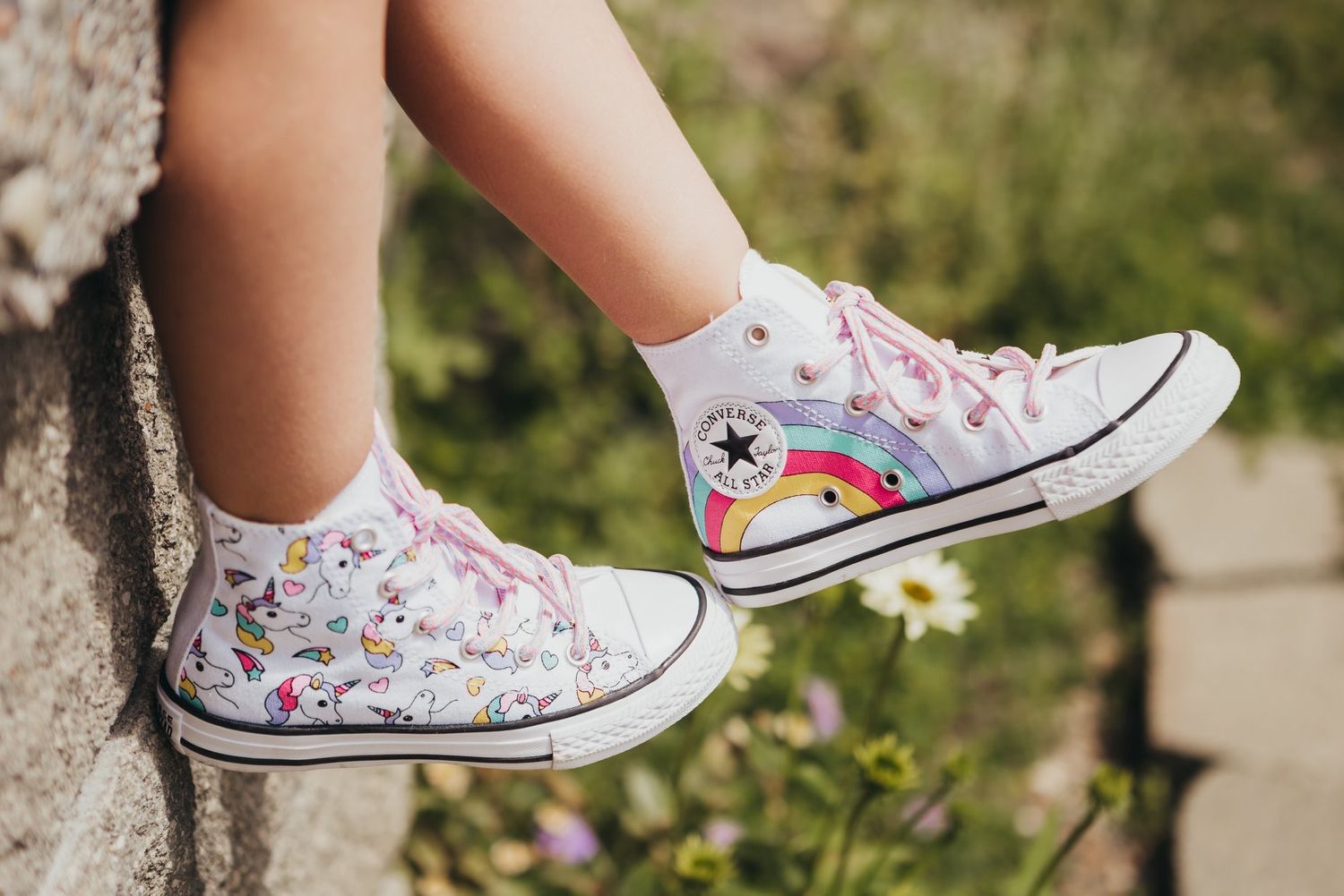 Child's feet wearing white Converse high-top sneakers with rainbow patch and pink laces outdoors.
