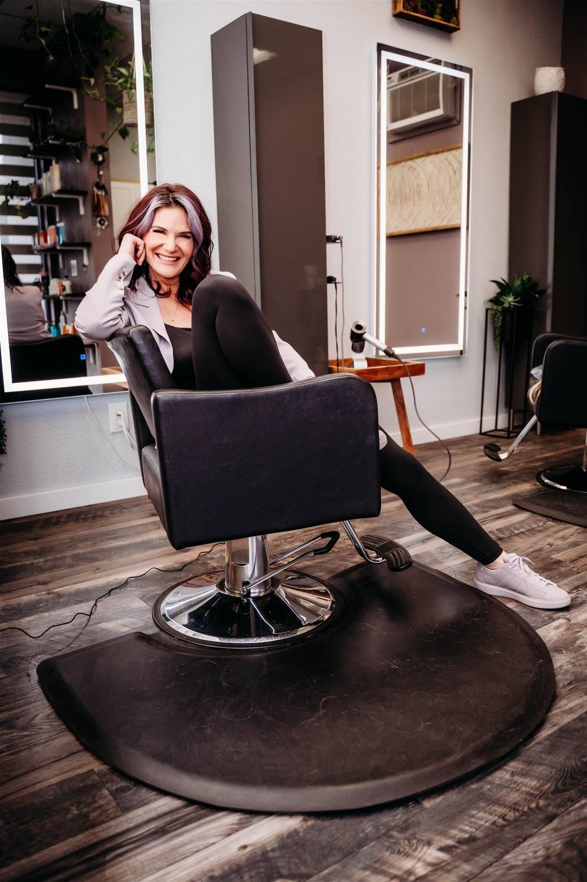 Smiling woman with dark hair sitting in a black salon chair in a modern hair salon interior.