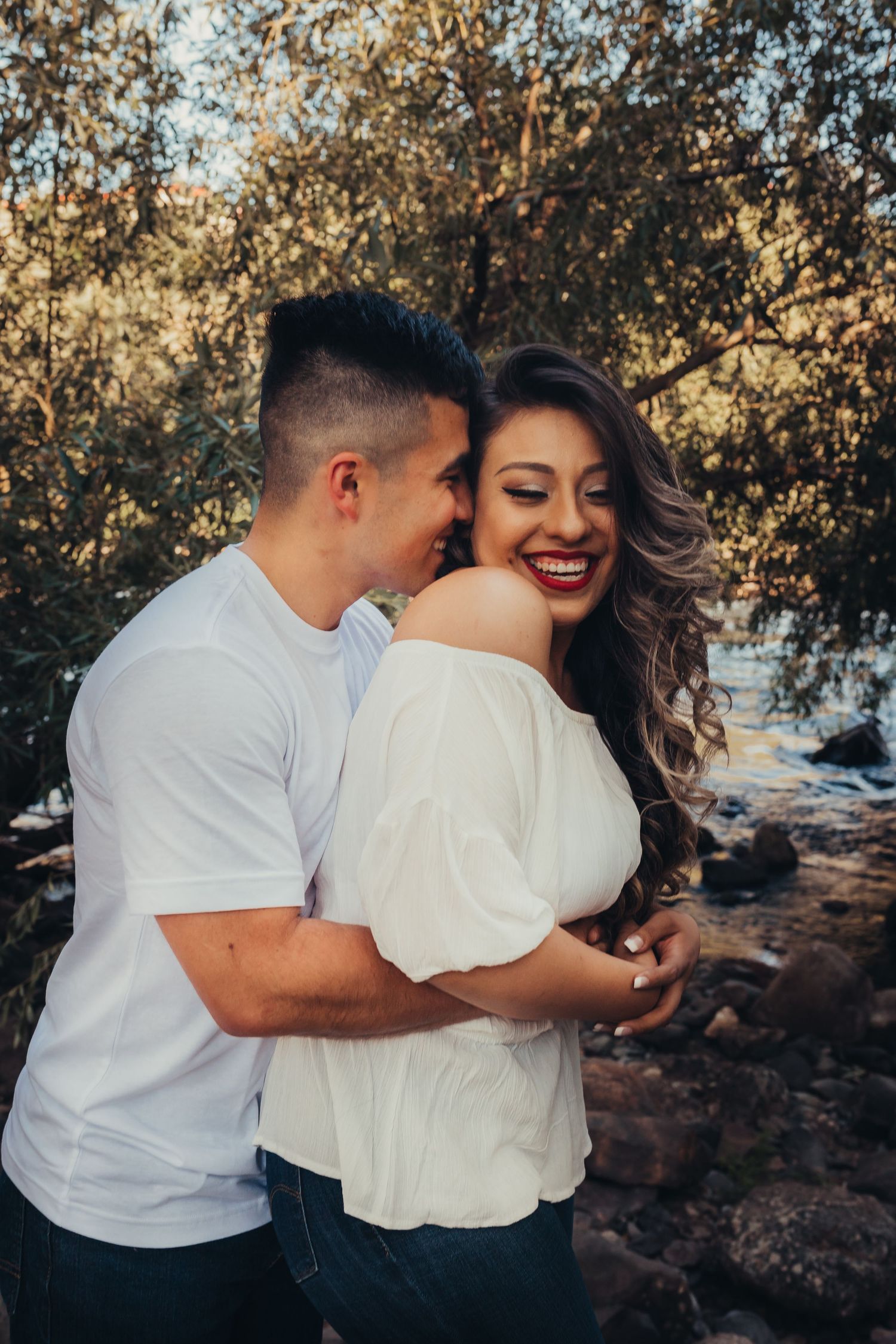 Happy couple embracing outdoors near trees, woman laughing joyfully in white off-shoulder top.