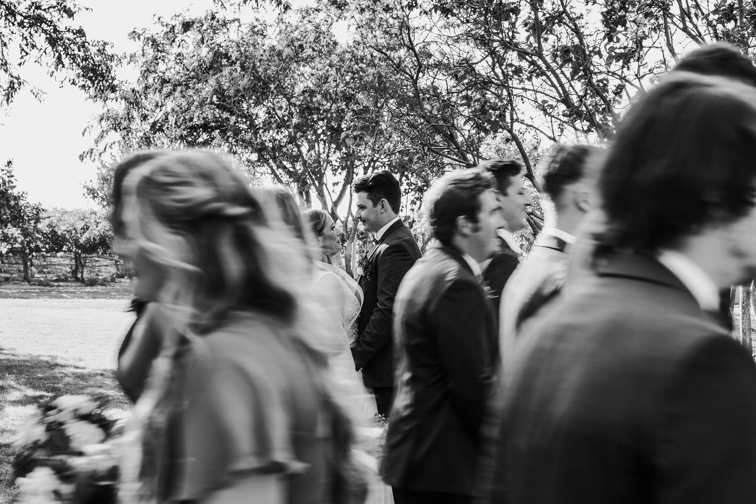 Black and white candid photo of wedding guests and groomsmen mingling outdoors under shady trees.