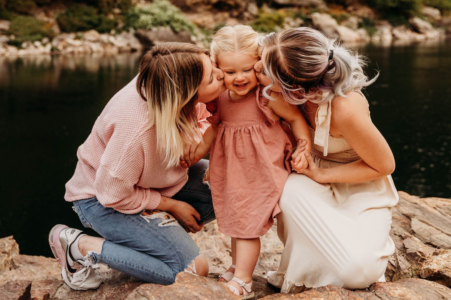 Two women kissing the cheeks of a laughing toddler girl in pink dress by a river.