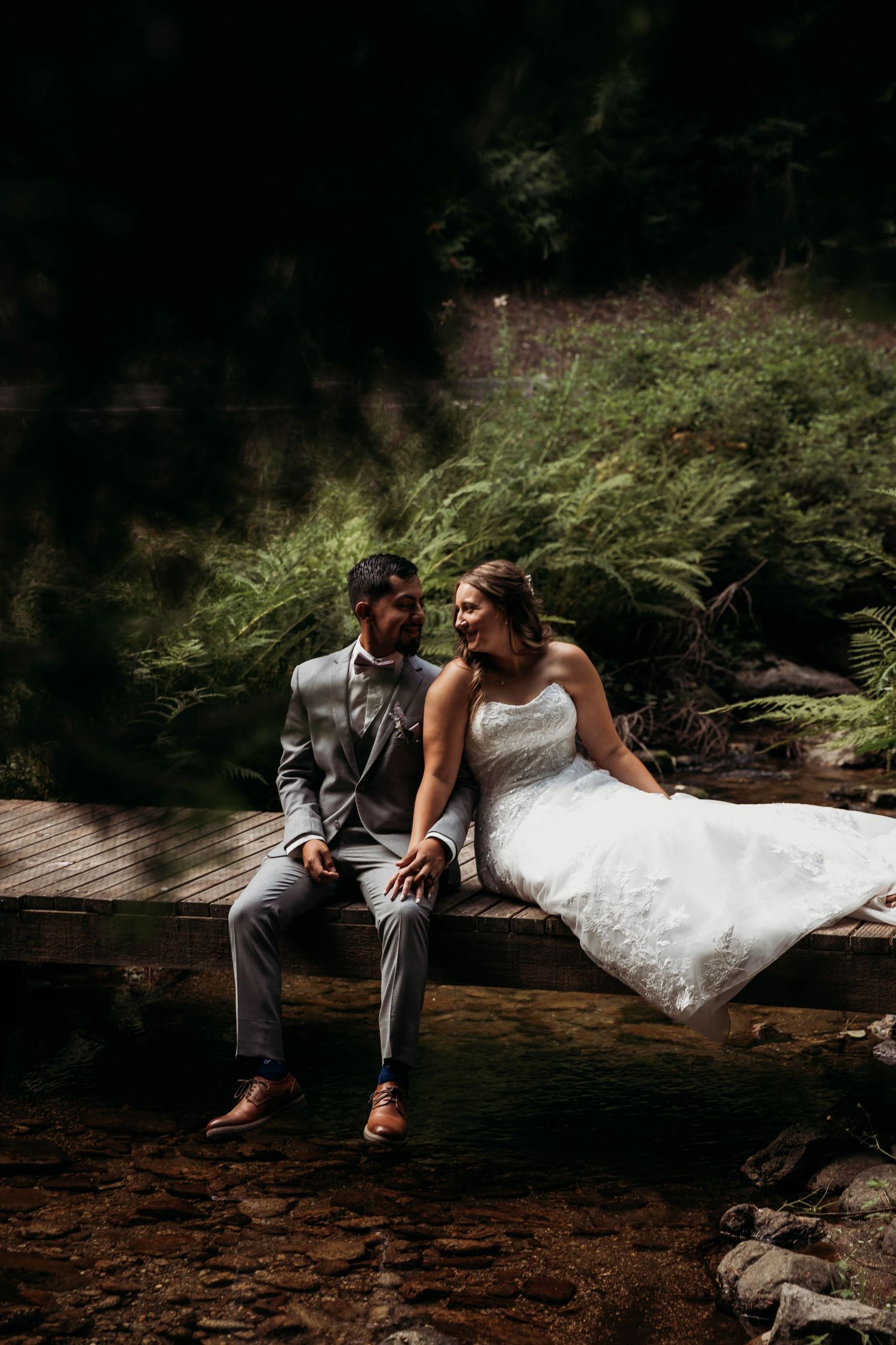 Bride and groom sitting together on a wooden bridge over a stream surrounded by lush green ferns in a forest.