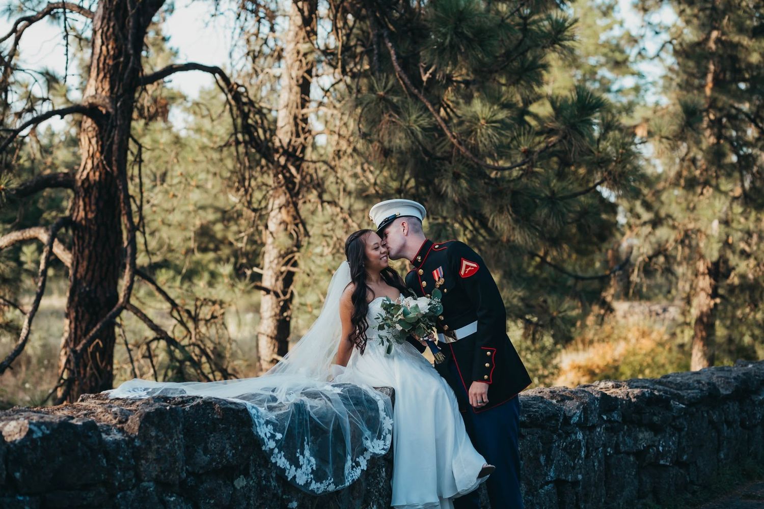 Marine groom in uniform kisses bride on a stone wall surrounded by pine trees.
