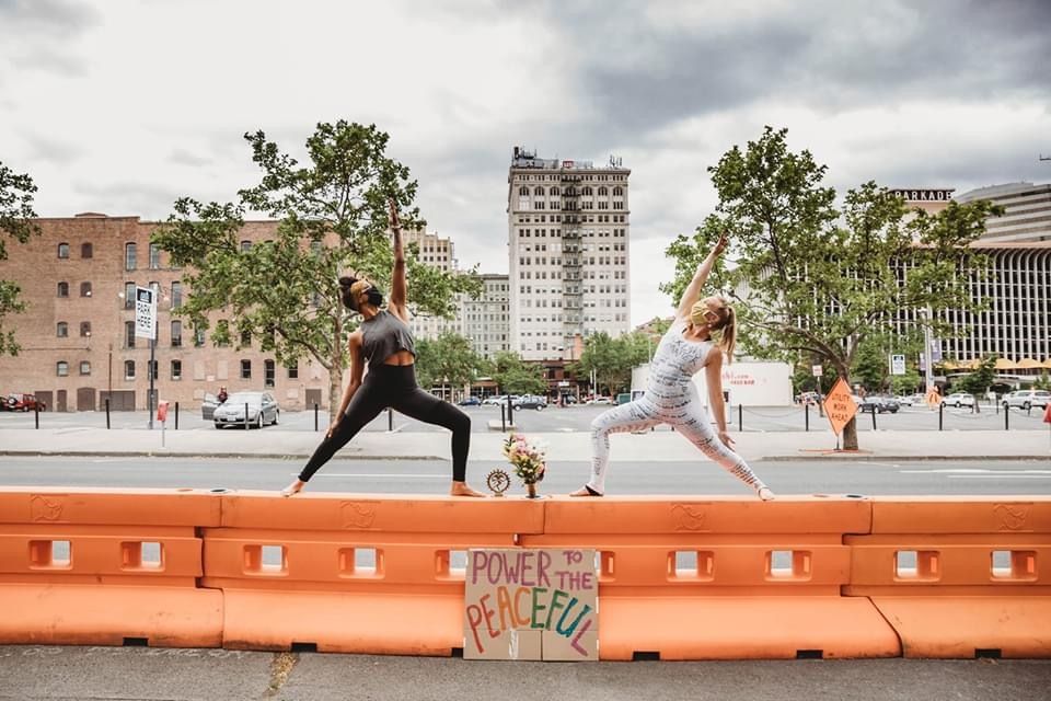 Two women doing yoga warrior poses on orange street barriers with a Power to the Peaceful sign outdoors.