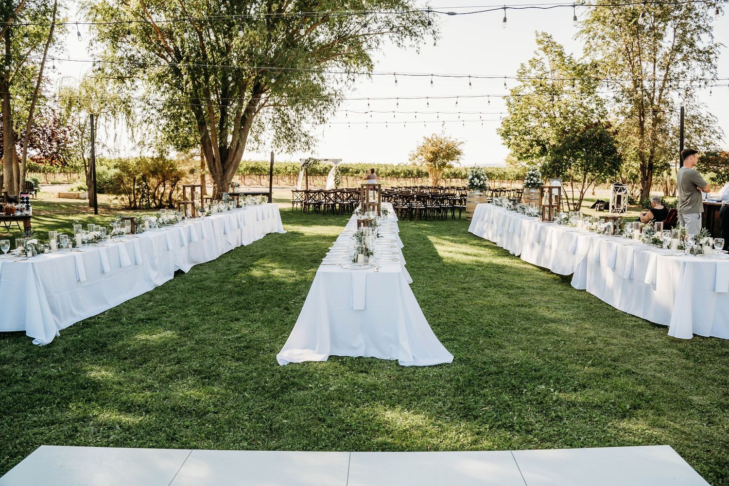 Outdoor wedding reception setup with long white tables, string lights, and lush green trees at golden hour.