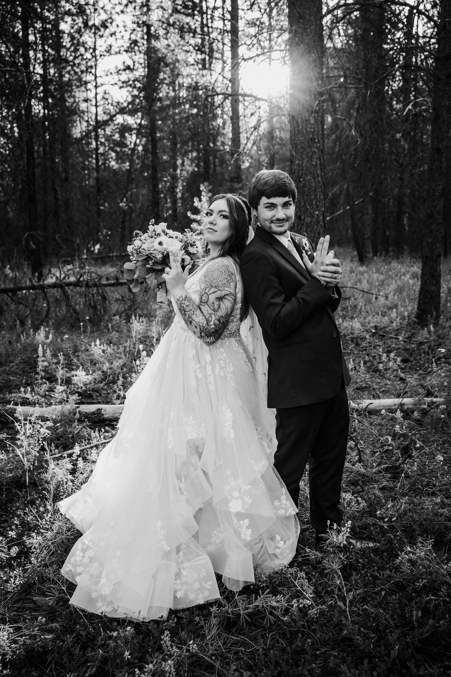 Black and white photo of bride with bouquet and groom embracing back-to-back in sunlit forest.
