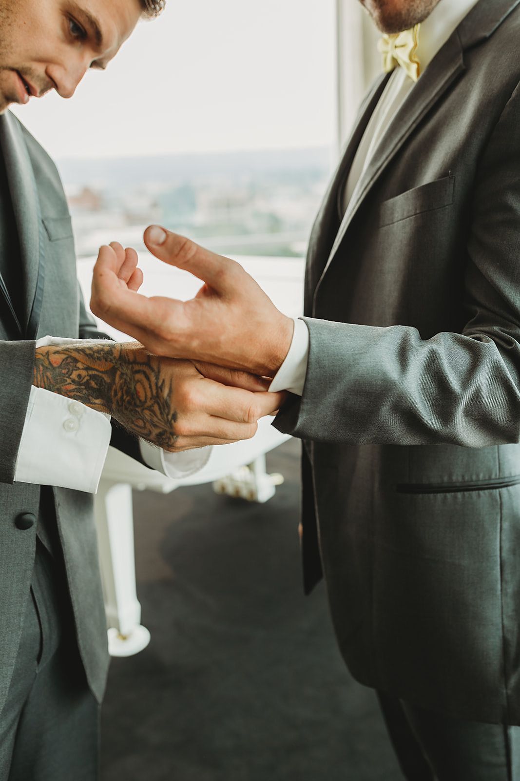 Two grooms in gray suits help each other with cufflinks before their wedding ceremony.