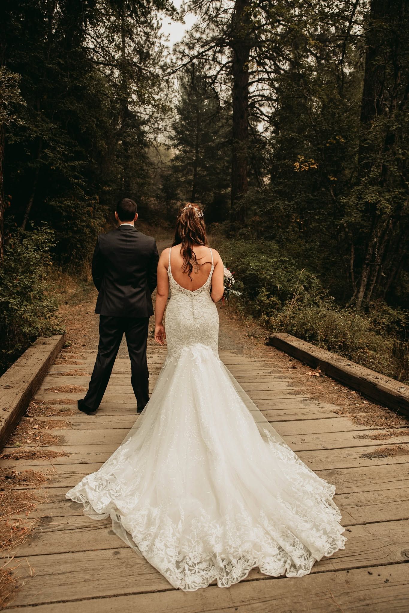 Bride in lace mermaid gown and groom in dark suit walk hand-in-hand on wooden path through forest.