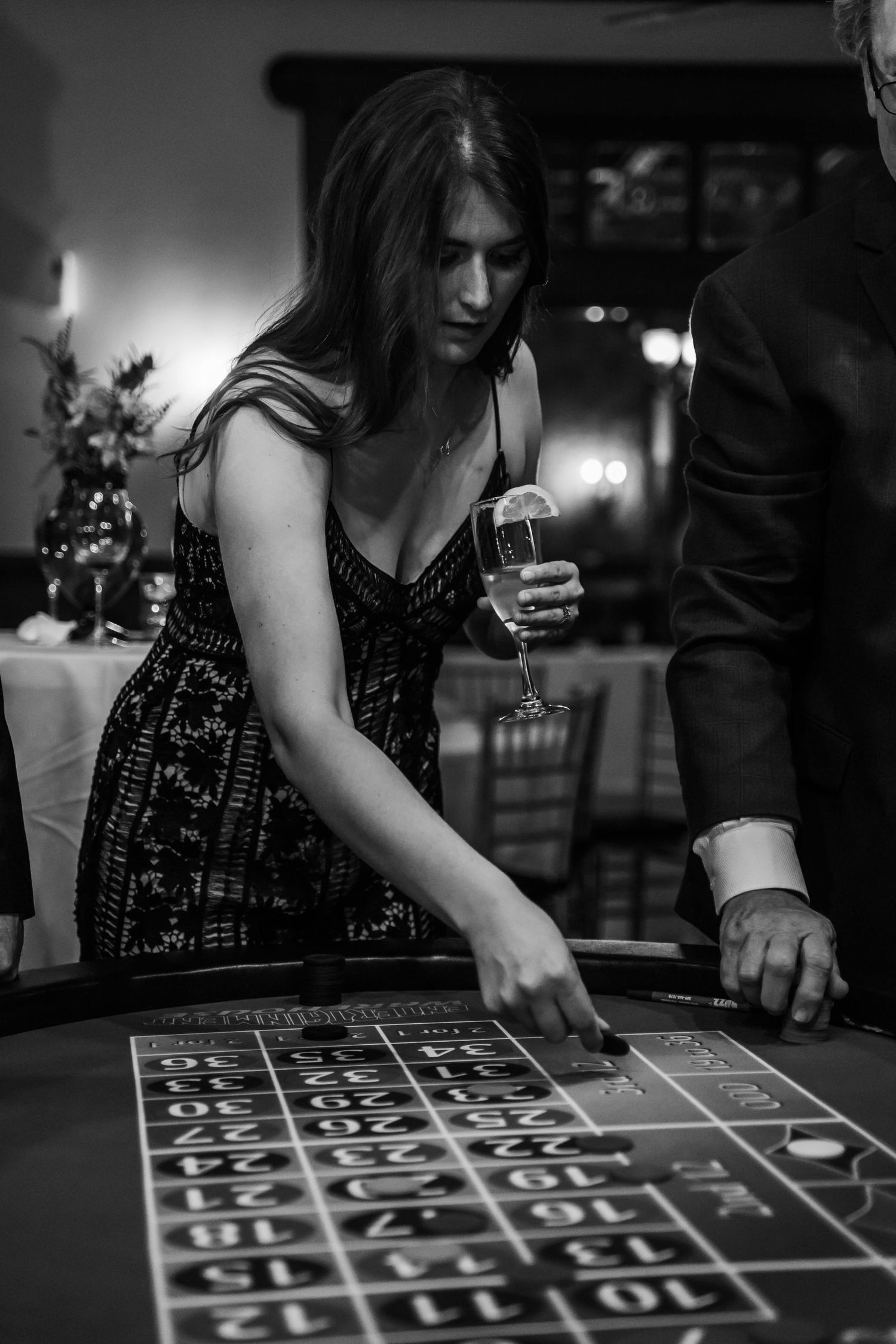 Black and white photo of a woman in a dress placing a bet at a roulette table holding champagne.