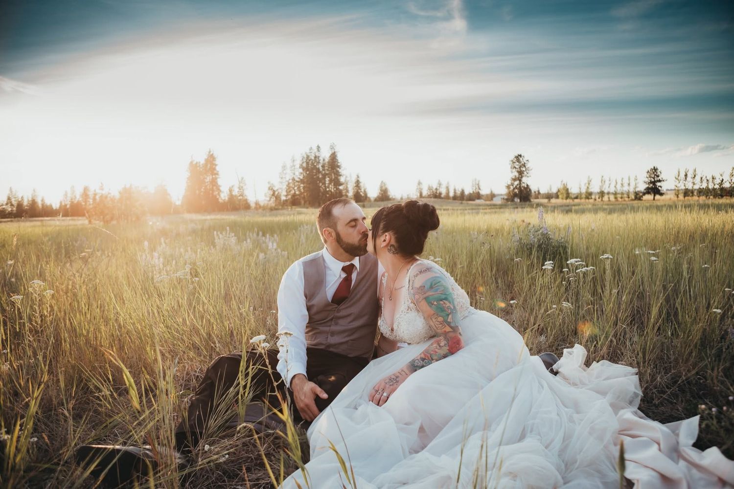 Bride and groom share a romantic kiss while sitting in a sunlit golden field on their wedding day.