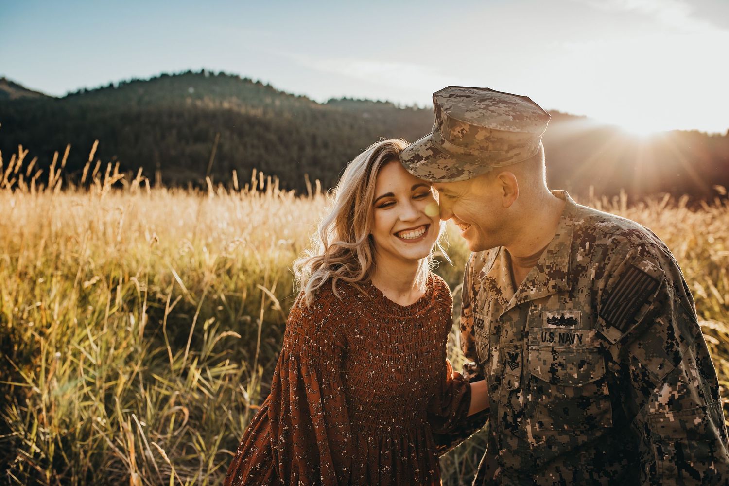 Smiling woman in rust dress and soldier in camouflage uniform share a tender moment in a golden field.