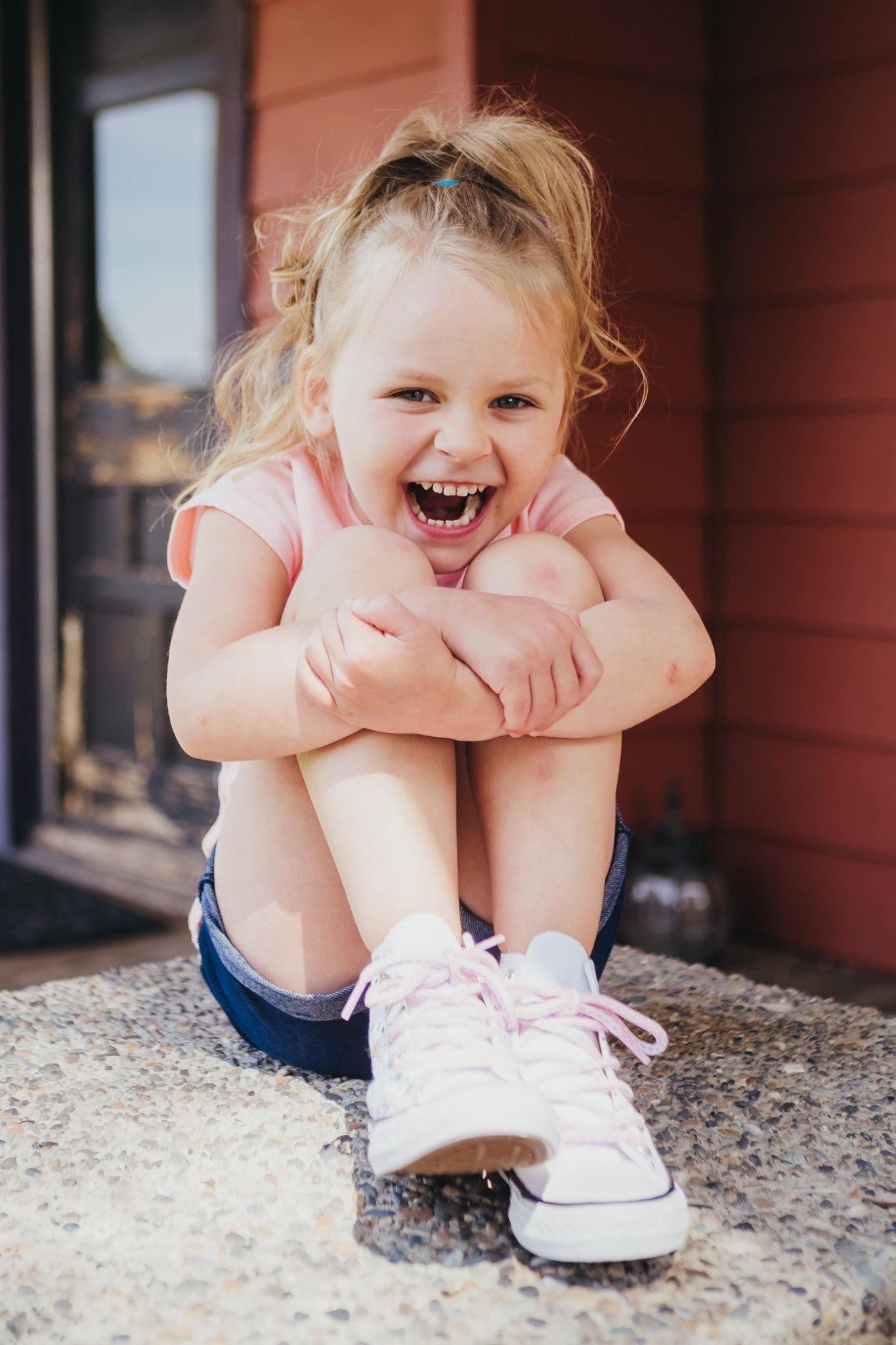 Happy laughing little girl in pink shirt and white sneakers sitting outdoors on a step.