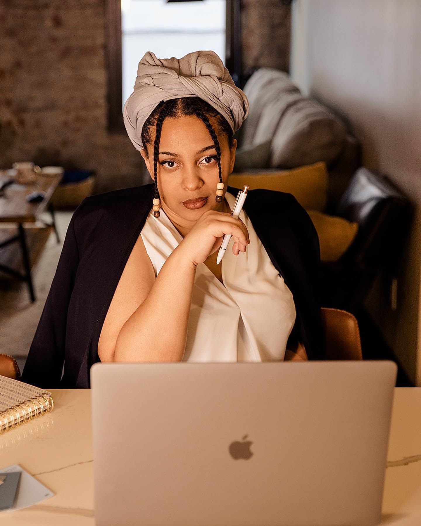 Professional Black woman in a head wrap sits thoughtfully at a desk with an Apple MacBook laptop.