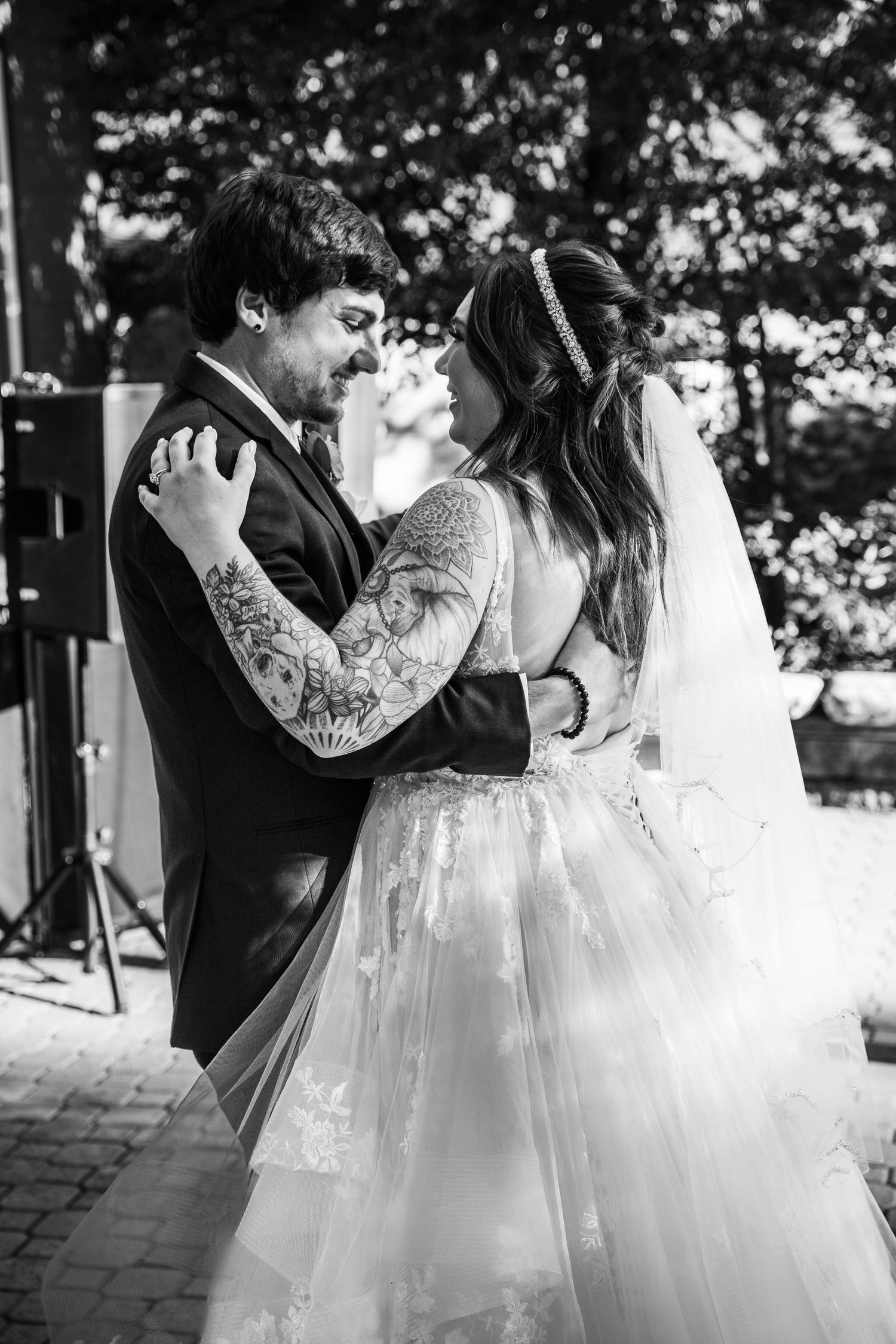 Black and white photo of tattooed groom and bride in lace veil sharing first dance at outdoor wedding.