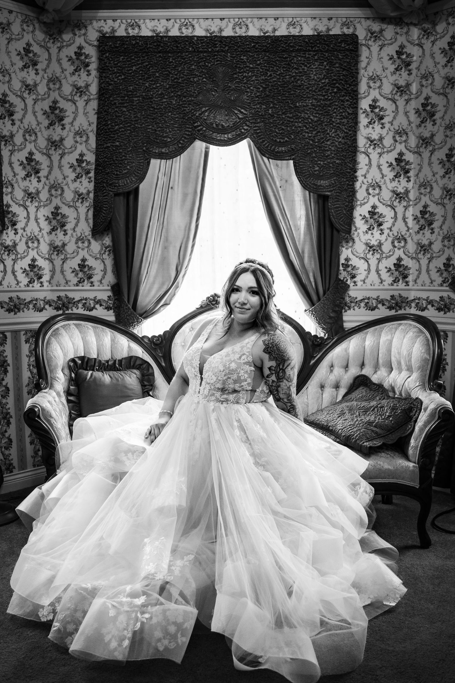 Bride in a voluminous ruffled wedding gown standing in an ornate vintage room with tufted sofas and draped curtains.