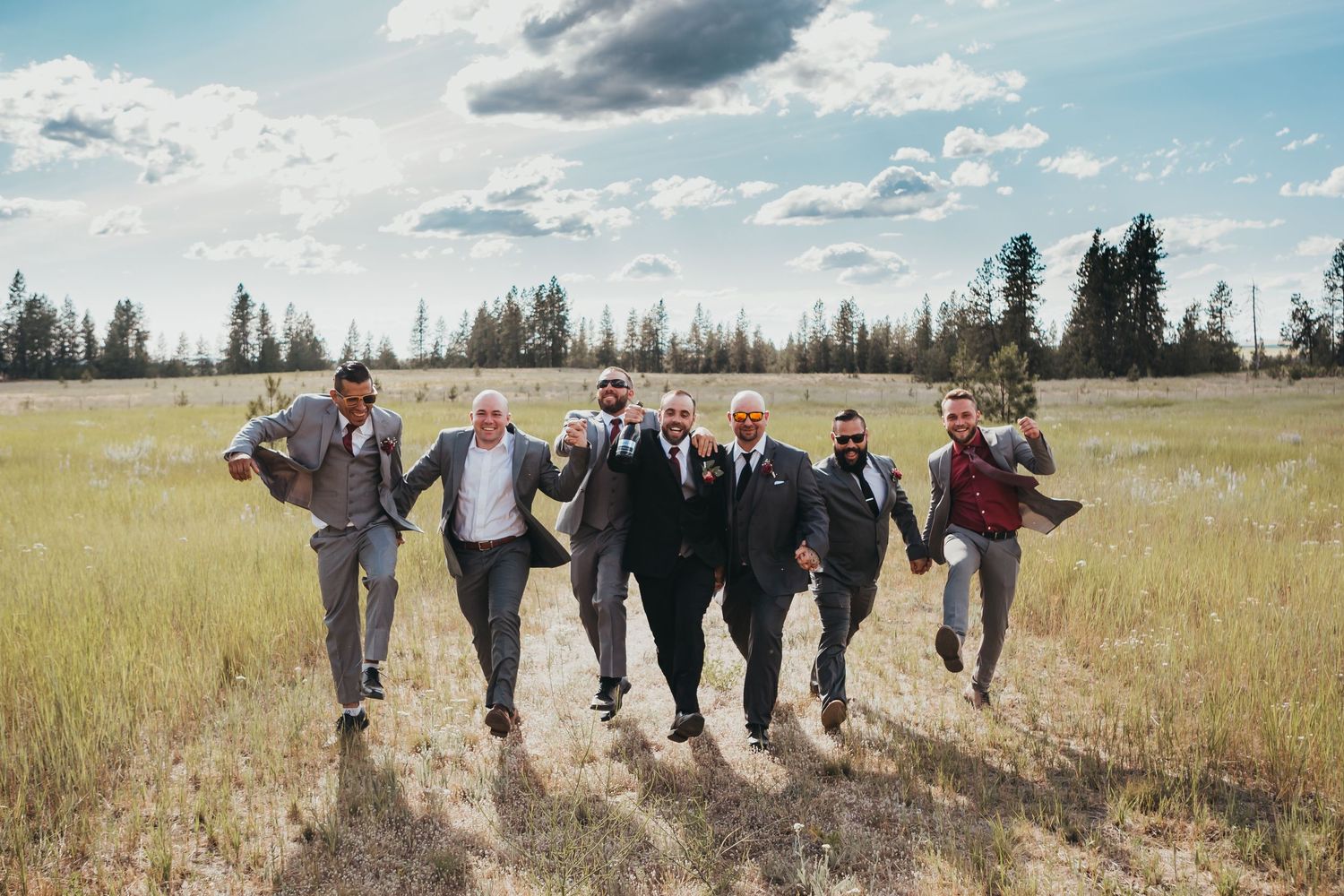Groom and groomsmen walk together laughing across a sunny meadow field in coordinating gray suits.