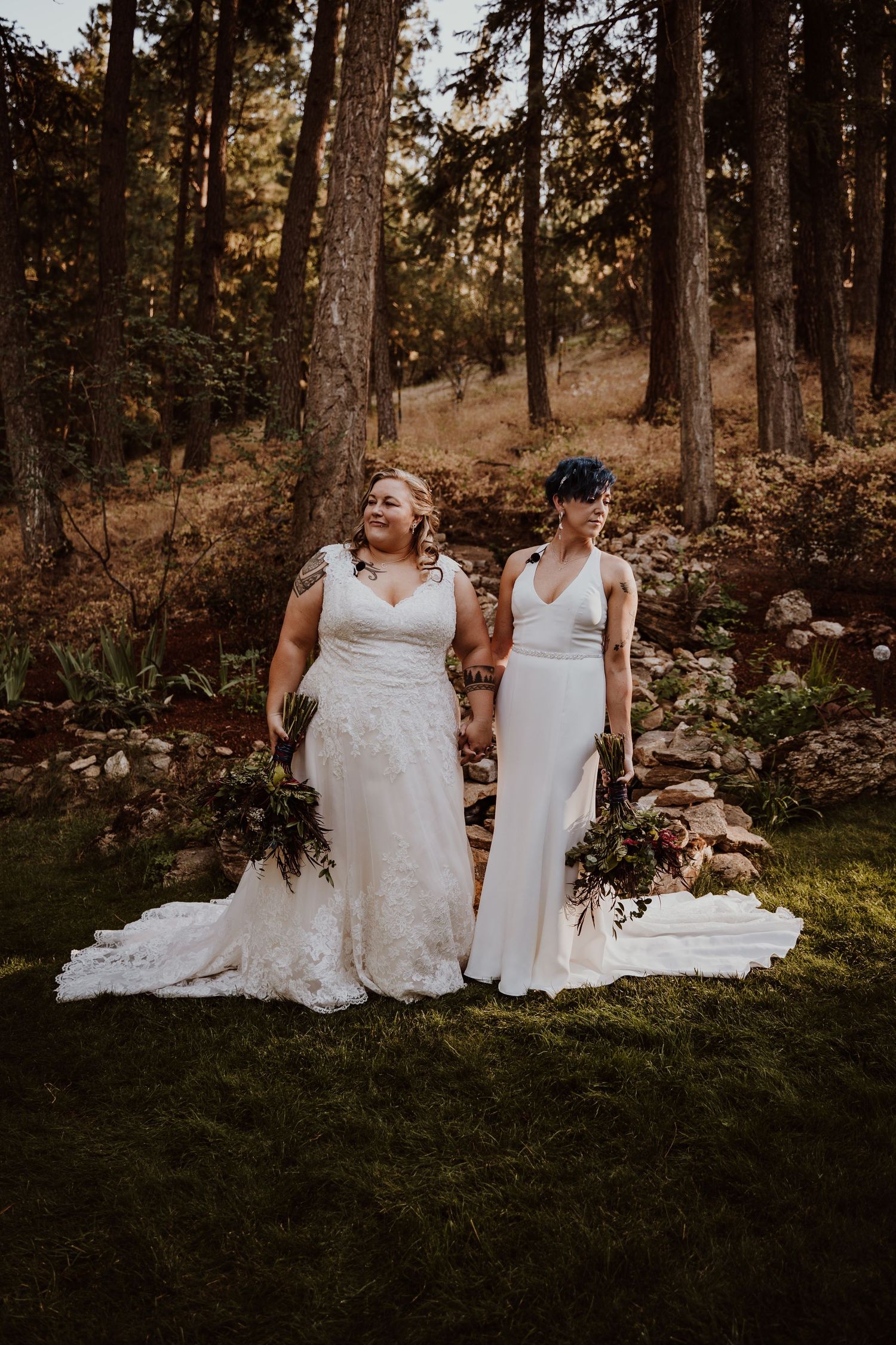 Two brides in white wedding gowns holding bouquets stand together in a forest setting.
