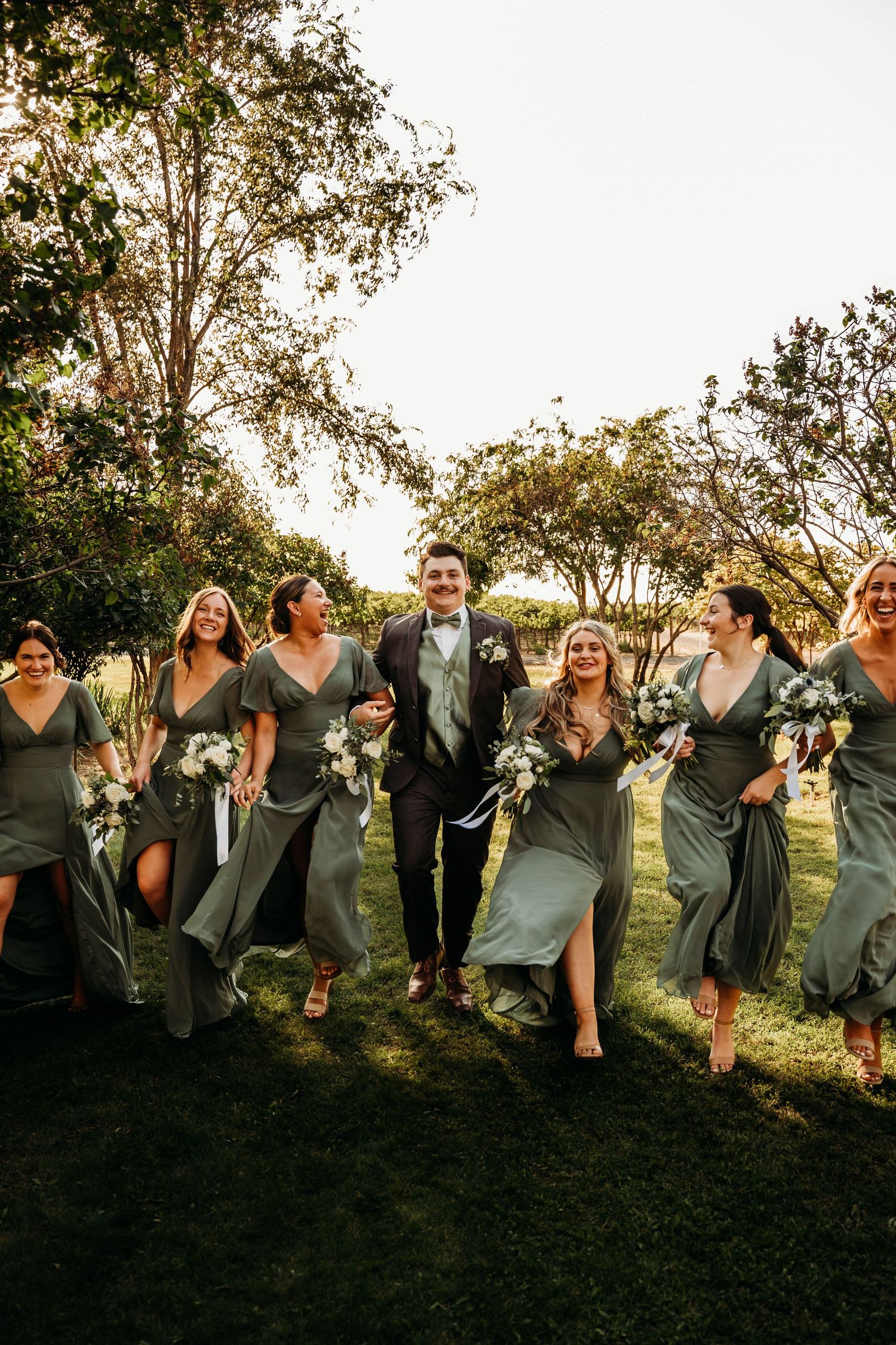 Groom walks joyfully with bridesmaids in sage green dresses carrying white bouquets in golden sunlight.
