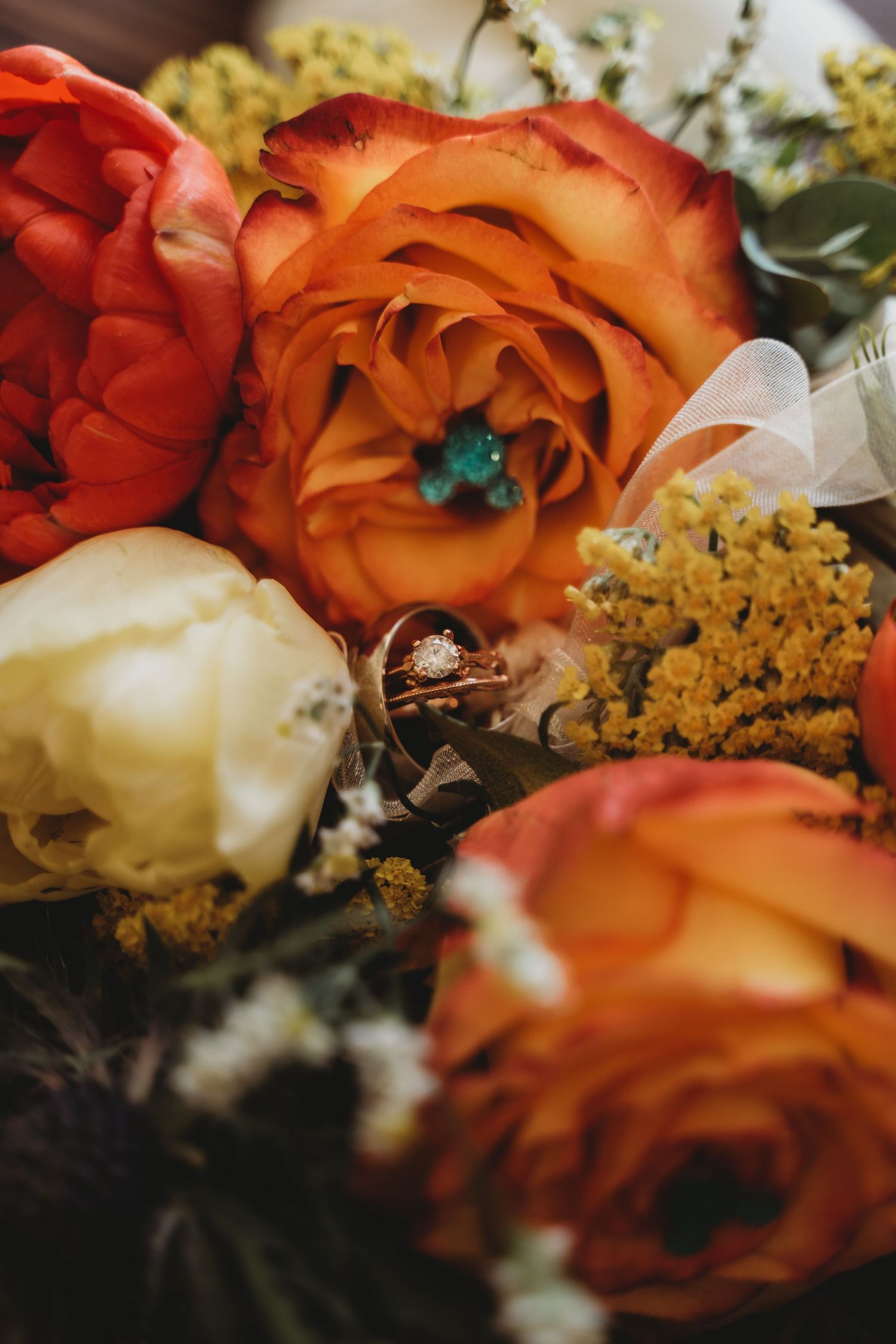Close-up of wedding rings nestled among orange and white roses in a bridal bouquet with yellow wildflowers.