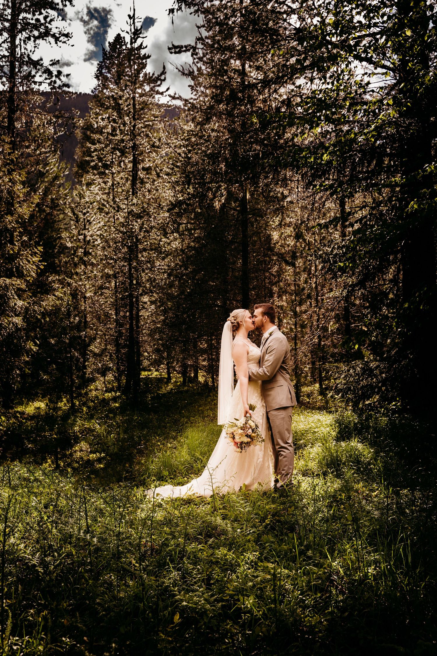 Bride and groom sharing a kiss in a sunlit forest clearing, bride in veil with bouquet.