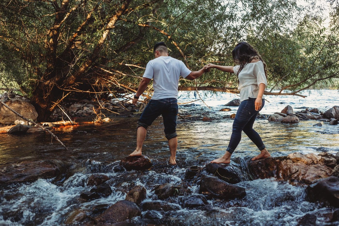 Couple holding hands while crossing rocky river stepping stones in lush forest.