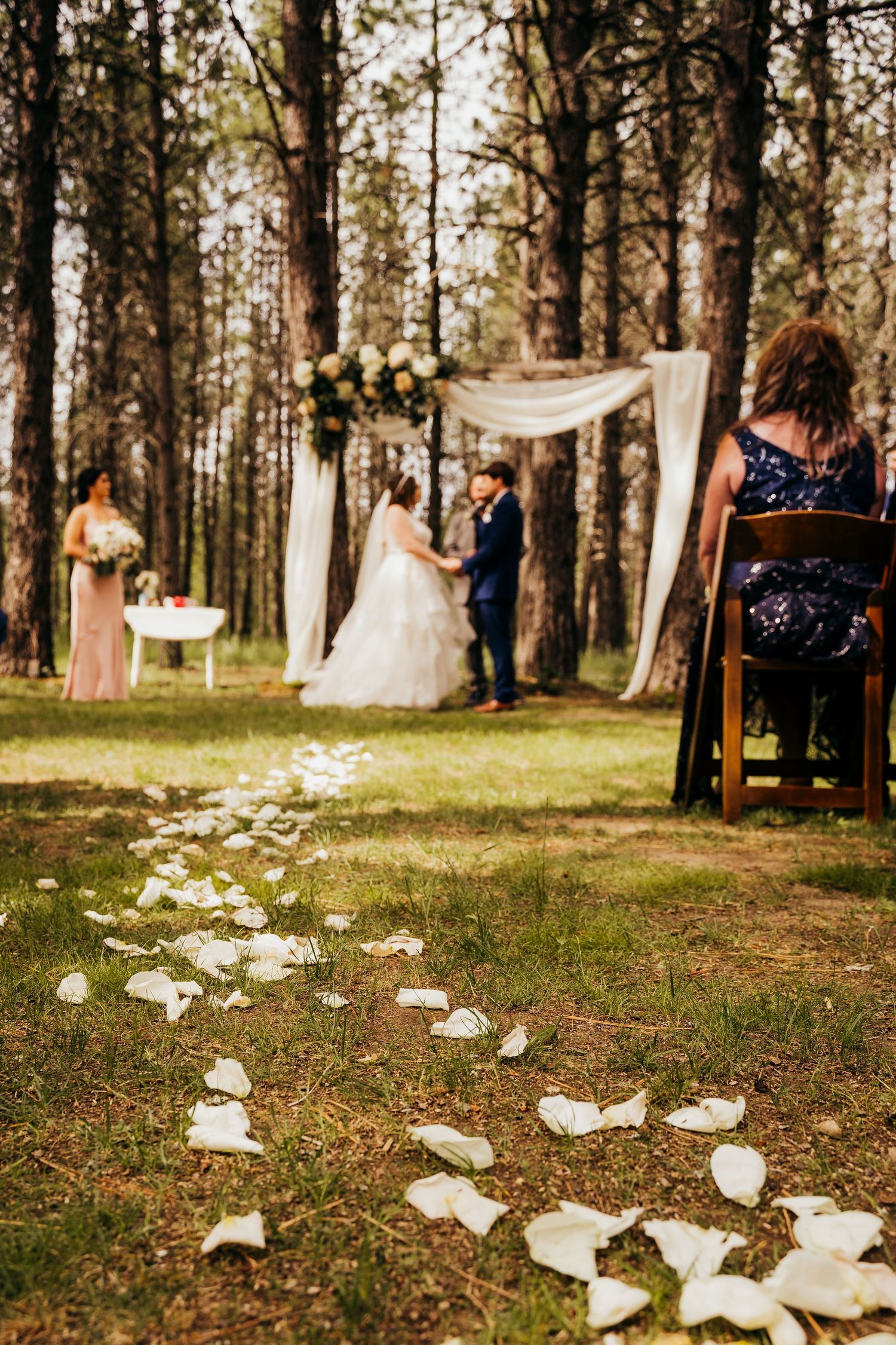 White rose petals line the aisle at an elegant outdoor forest wedding ceremony.