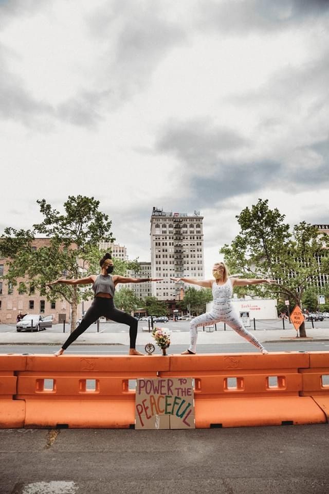 Two women performing warrior yoga poses on orange barriers with a Power to the Peaceful sign in a city.