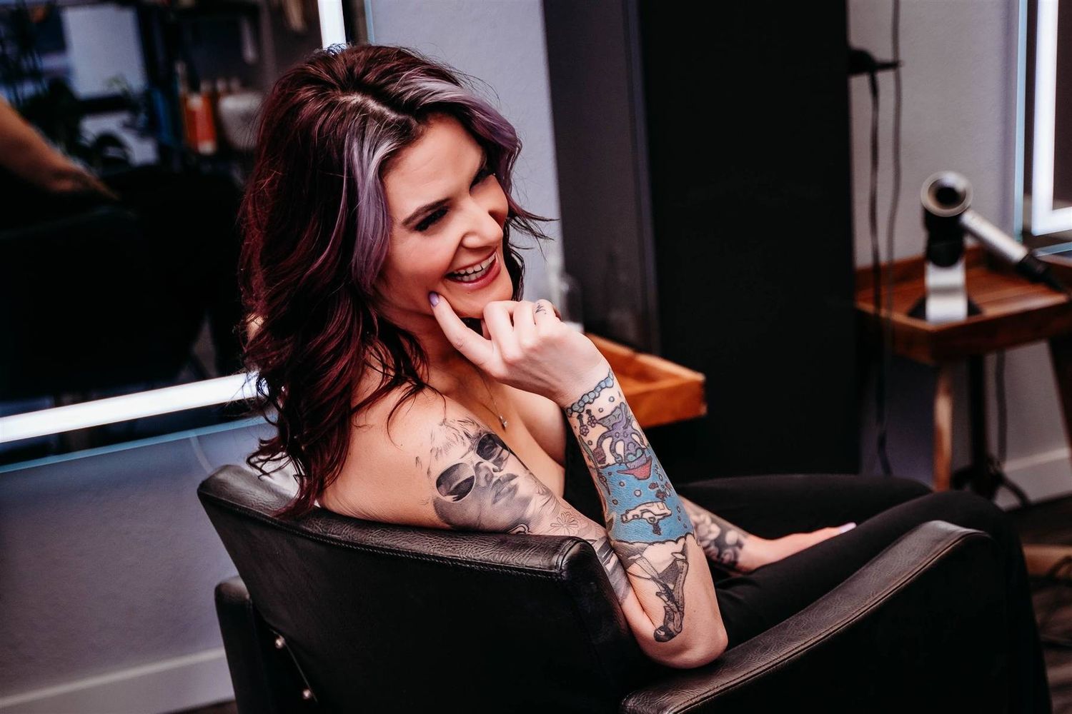 Tattooed woman with dark wavy hair laughing while sitting in a salon chair, talking on the phone.