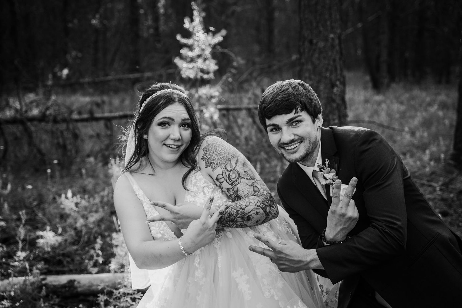 Black and white photo of smiling bride and groom making fun hand gestures during forest wedding shoot.