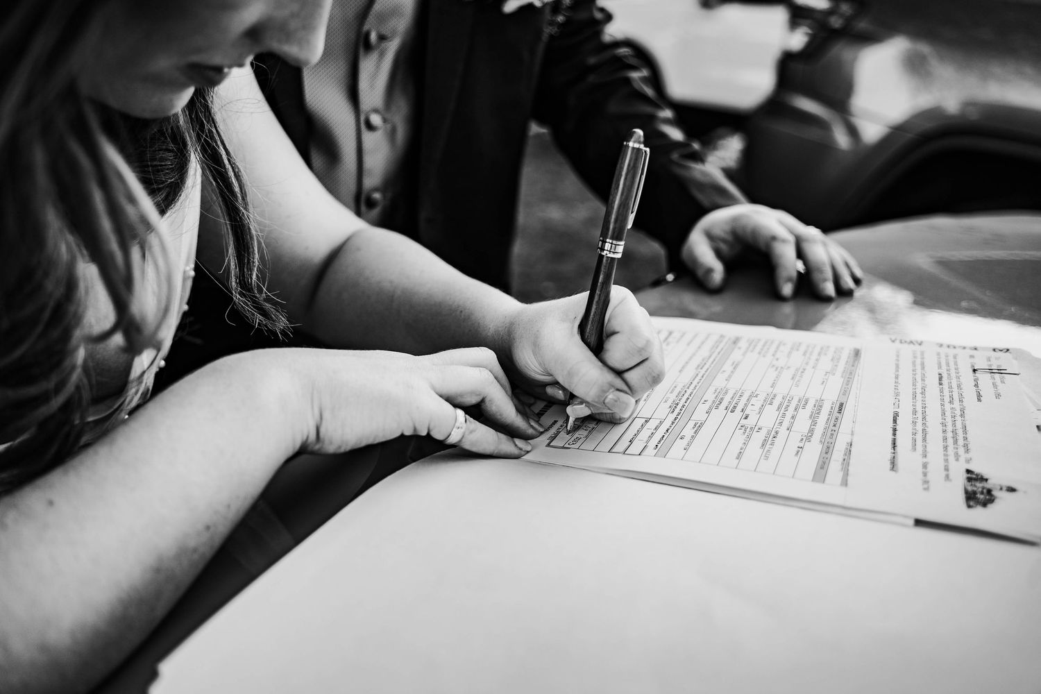 Black and white close-up of bride signing wedding marriage license document with a pen at a table.