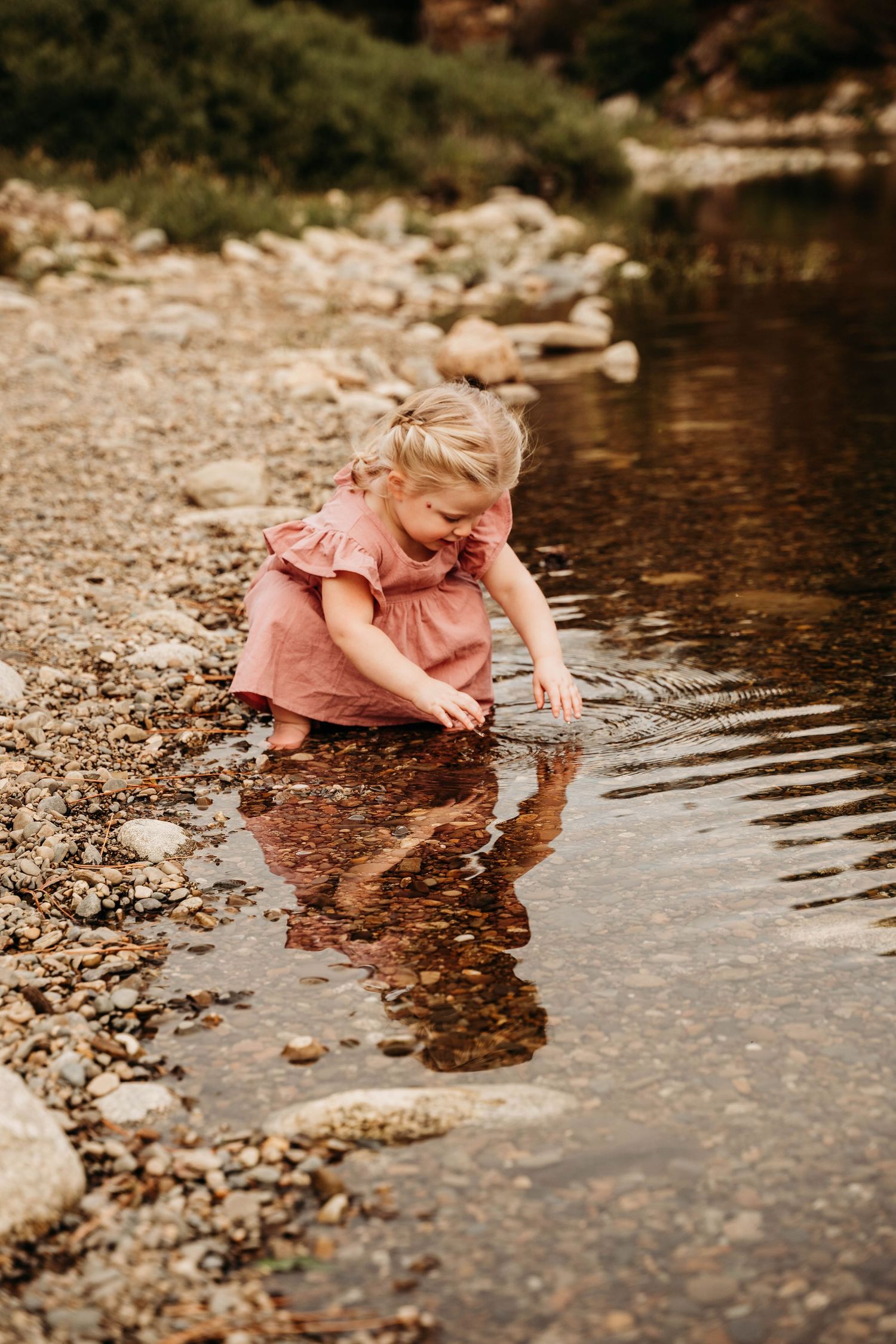 A young toddler in a pink dress crouches by a rocky shoreline, playing in the shallow water with her hands.