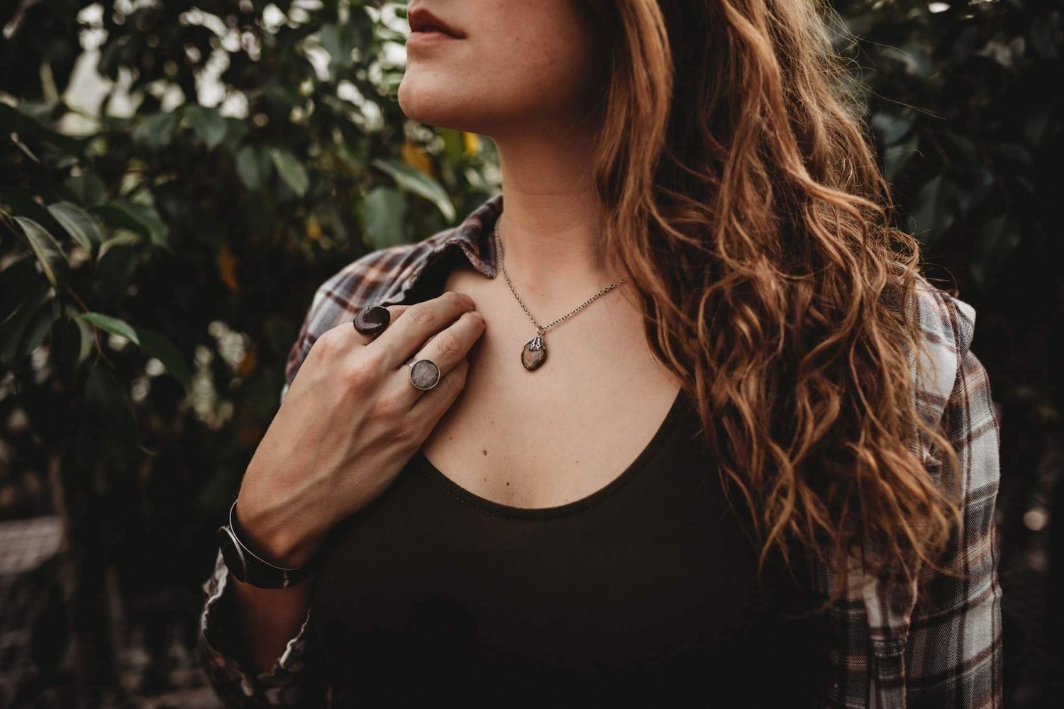 Woman in plaid shirt and black top displays layered necklaces and ring among green foliage.