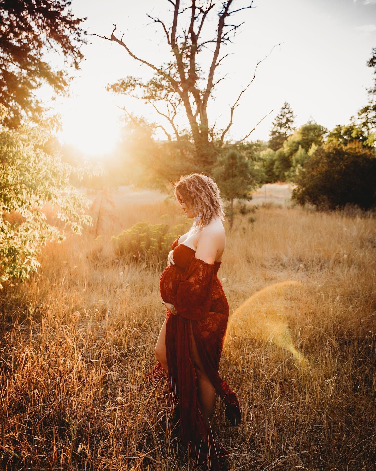 Pregnant woman in burgundy gown standing in golden field during stunning sunset.