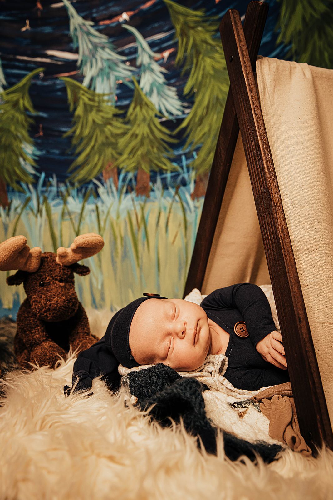 Newborn baby in black outfit sleeping beside moose stuffed animal under a teepee tent prop.